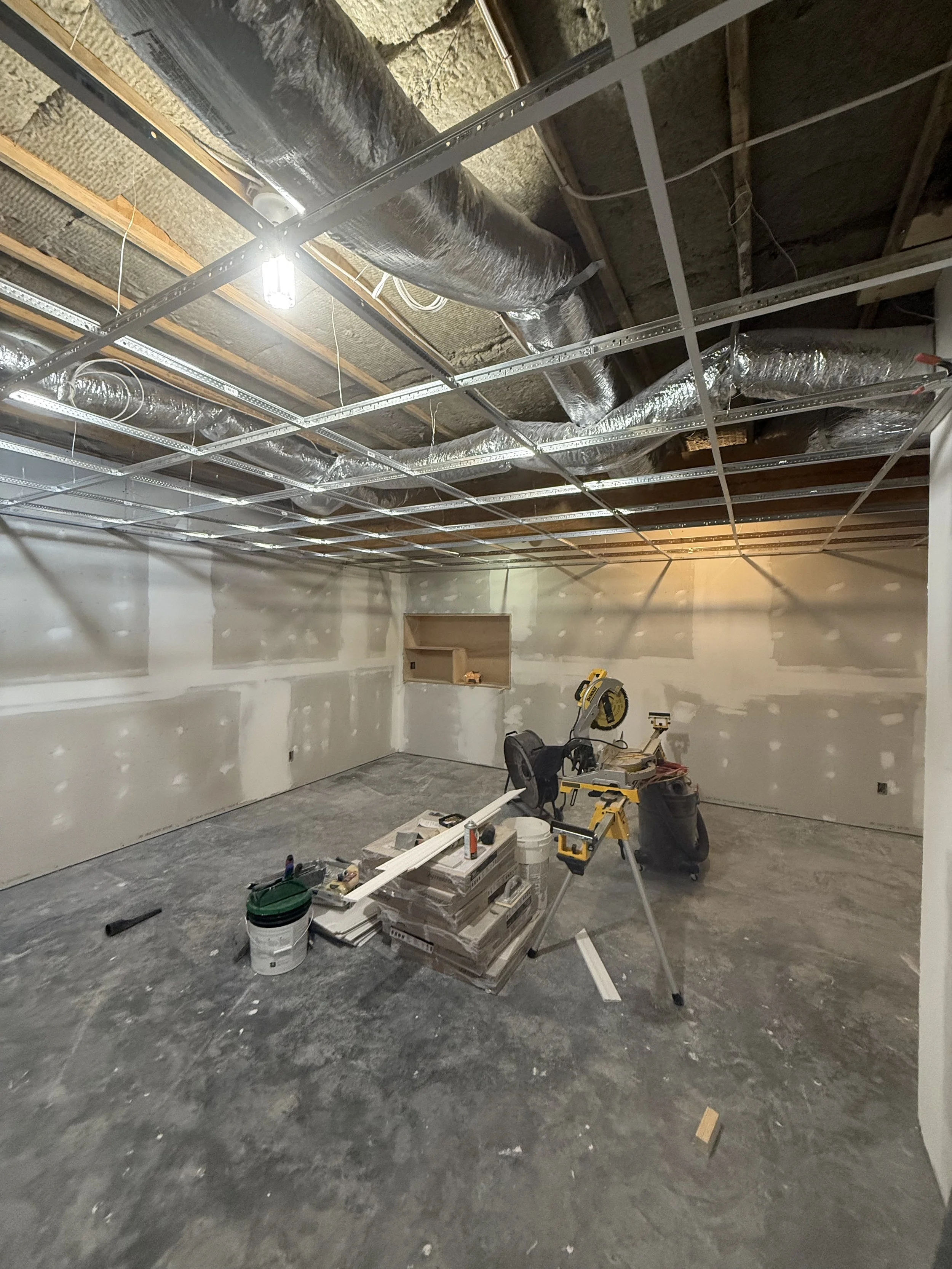 Interior of a room under construction or renovation with drywall walls and a concrete floor. The ceiling framework is visible with metal studs and ductwork. Construction tools and supplies are scattered on the floor, including a miter saw, buckets, and boxes.