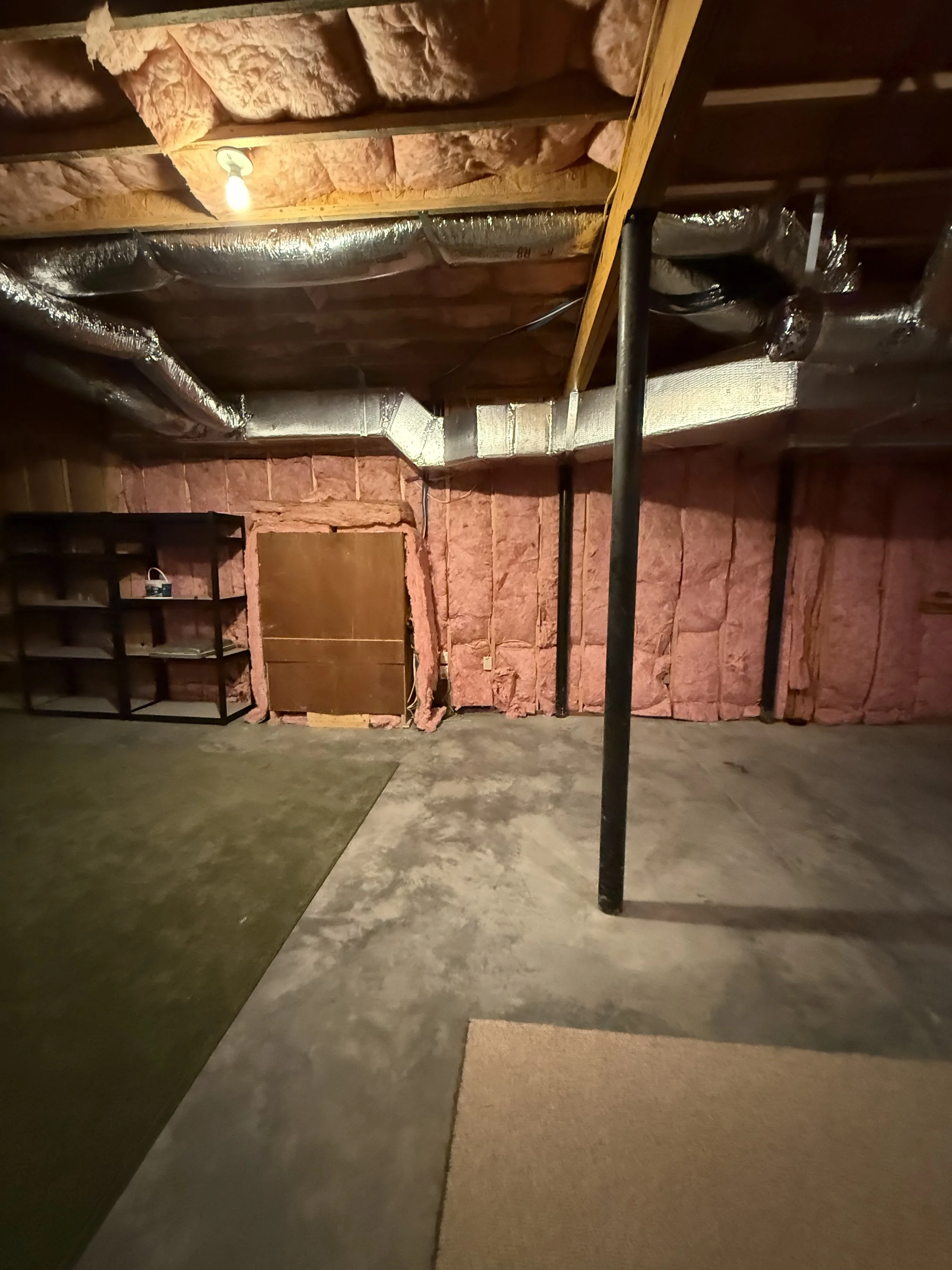 A basement with insulation, ductwork, a single ceiling light, a black shelf unit, and two different colored rugs on the concrete floor.