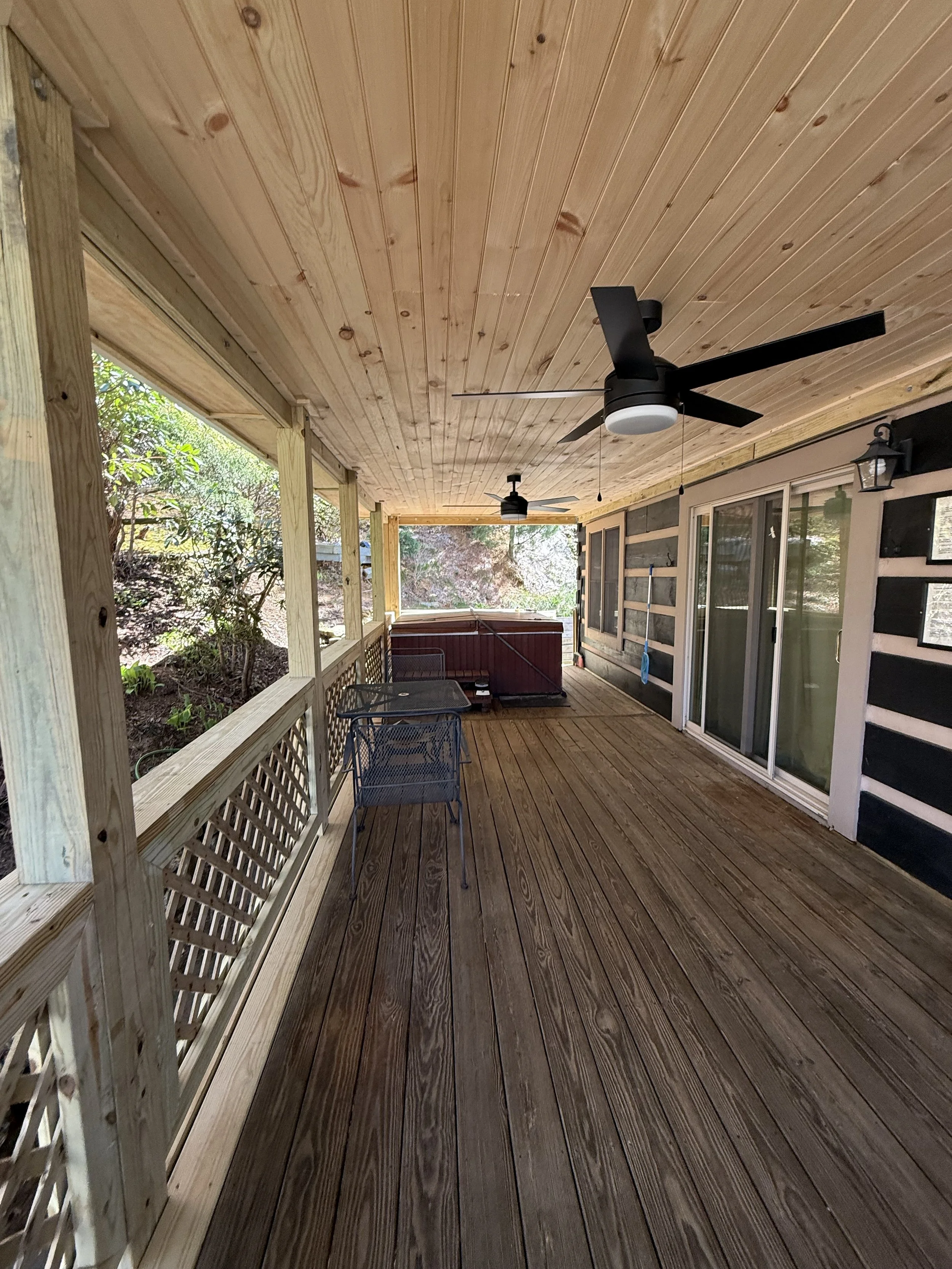 Wooden porch with ceiling fans and outdoor furniture, overlooks a garden with trees and bushes.