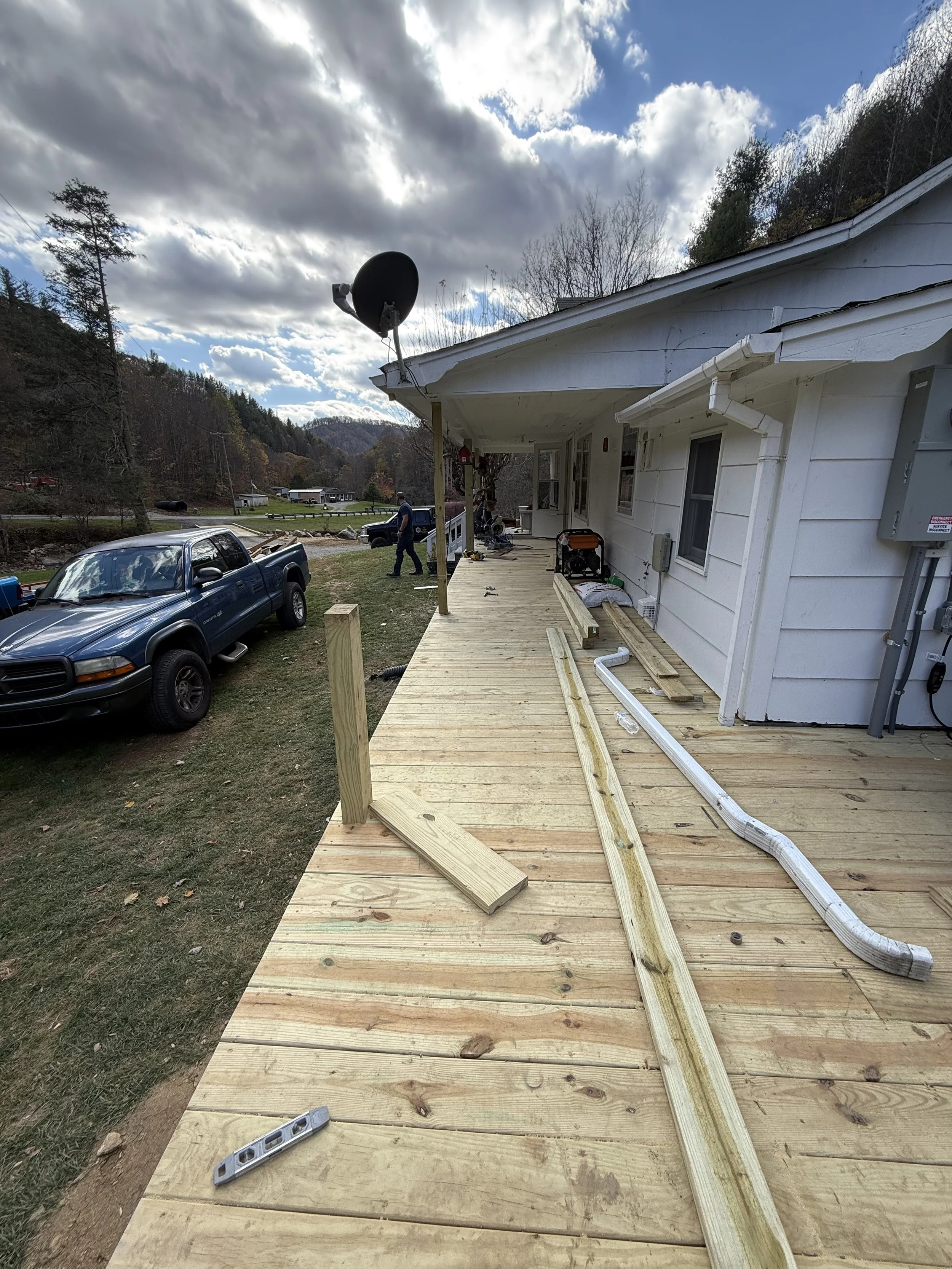 Partially built wooden deck attached to a white house, with construction tools and materials on it. Cars parked on the grass, with a background of trees and cloudy sky.