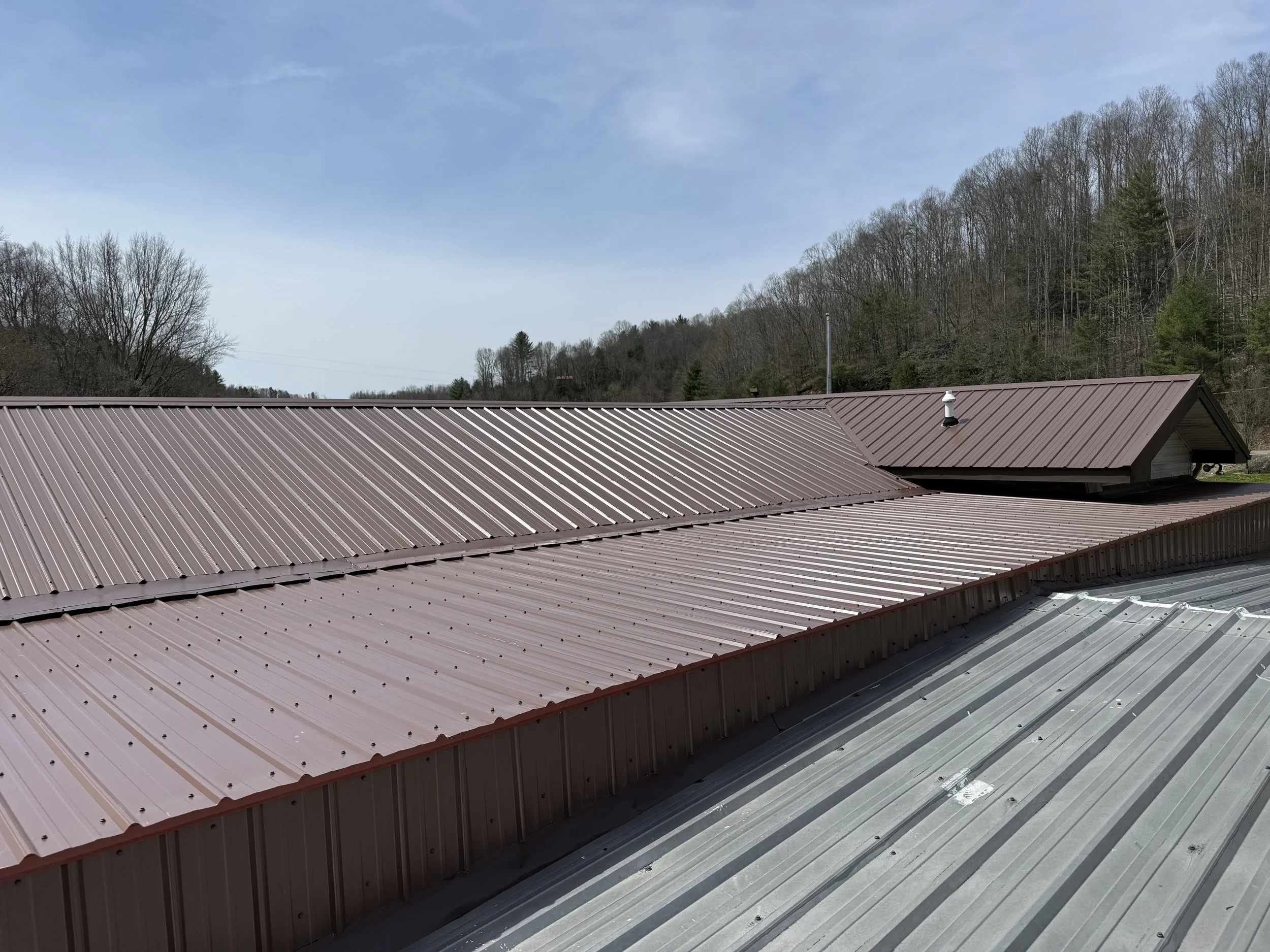 Metal roof with brown panels in the foreground, a blue sky with some clouds, and a wooded hillside in the background.
