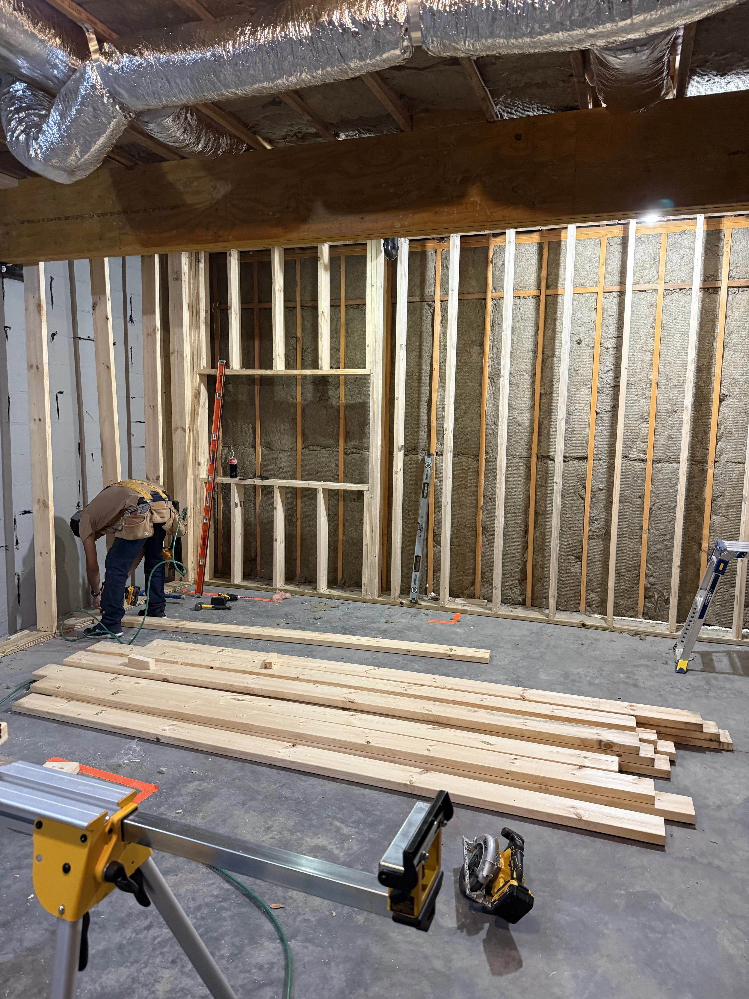 Construction worker assembling wooden framing in an unfinished room with exposed insulation and ceiling ducts, various tools and lumber scattered on the floor.