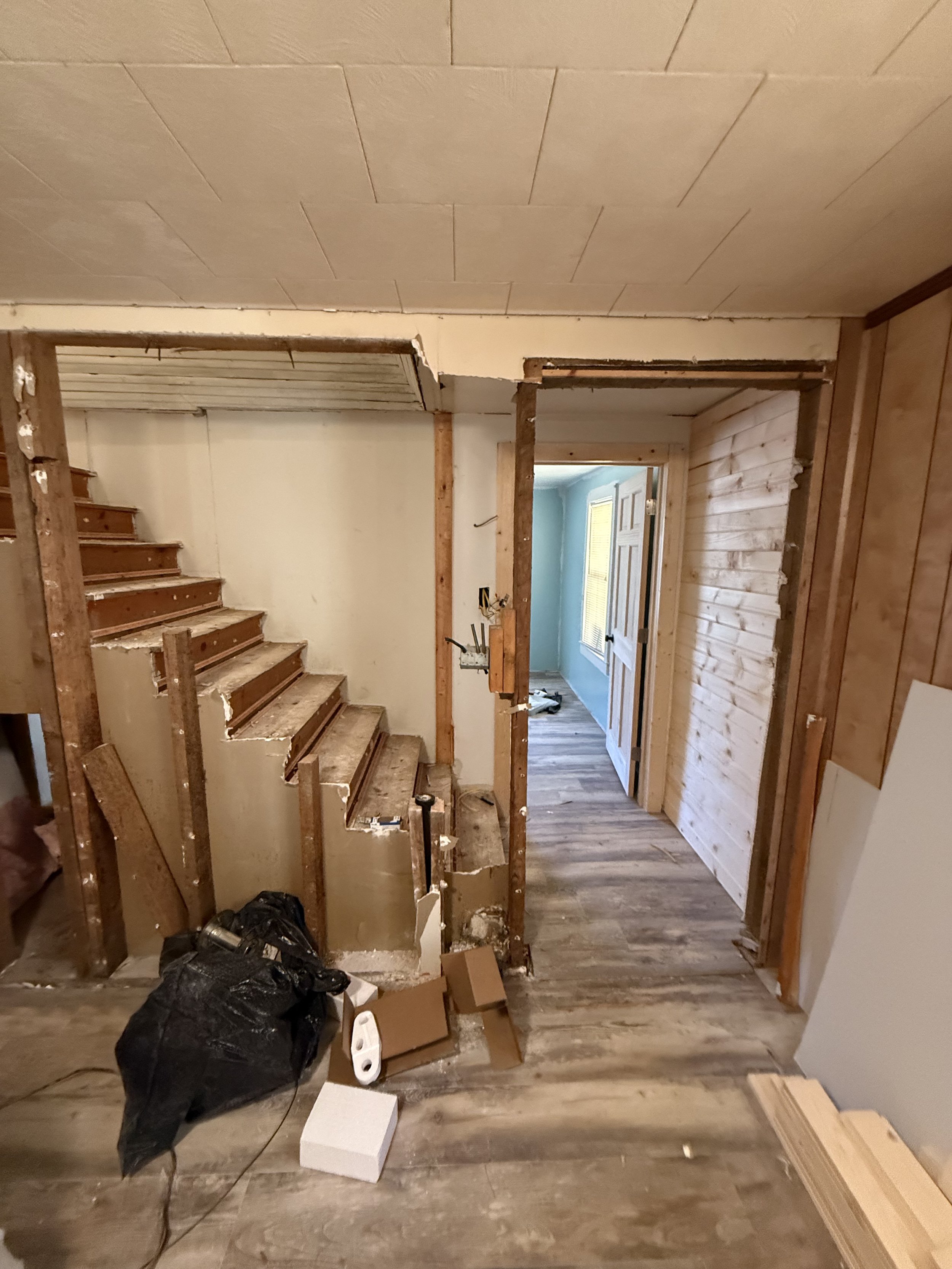 Interior of a house under renovation showing exposed wooden framing for a wall or doorway, with construction tools, a black trash bag, and debris on the floor. Part of a staircase and hallway is visible in the background.