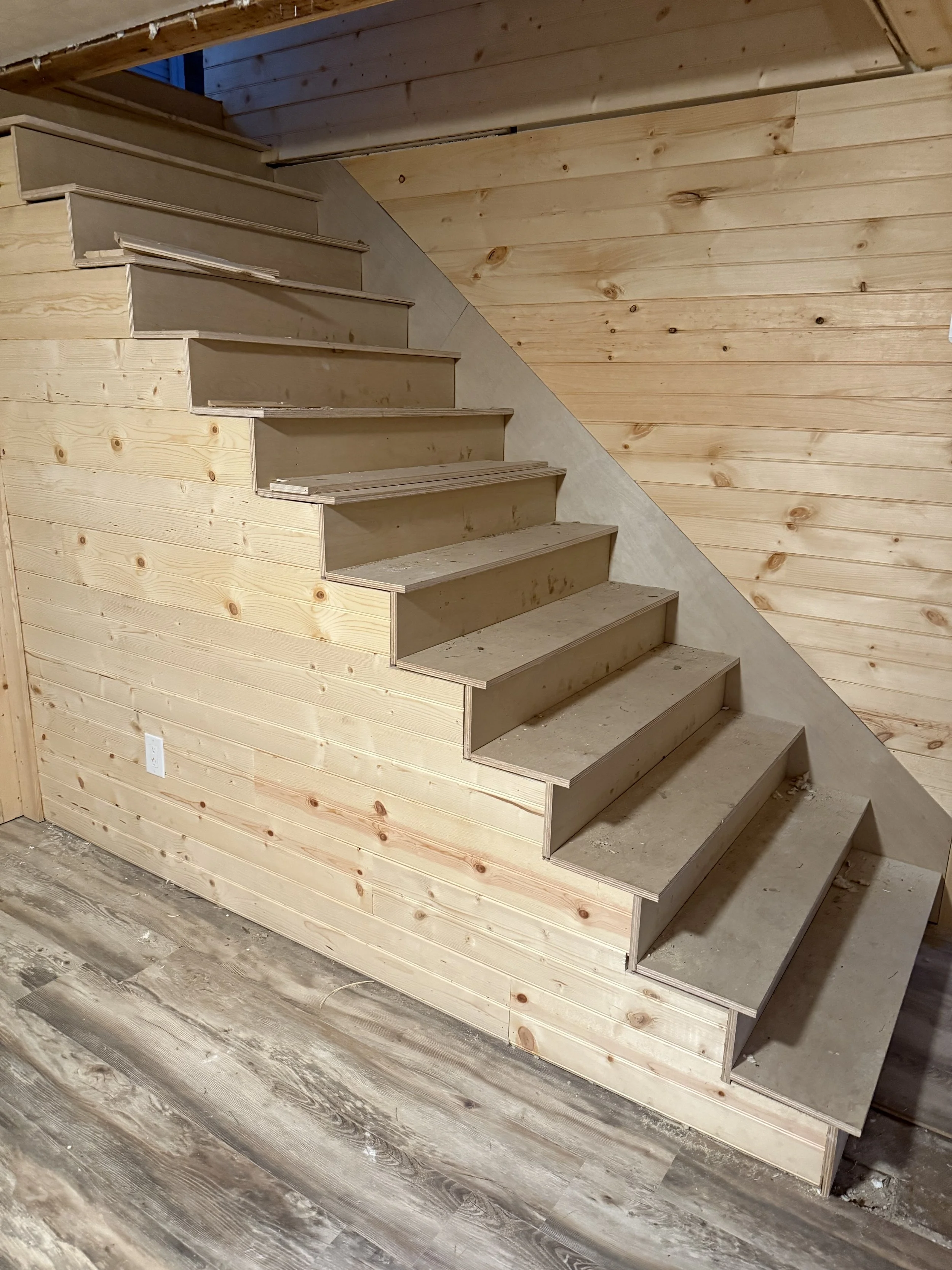 Unfinished wooden staircase with light-colored wood panels on the walls and floor, and concrete on the staircase steps in a construction area.