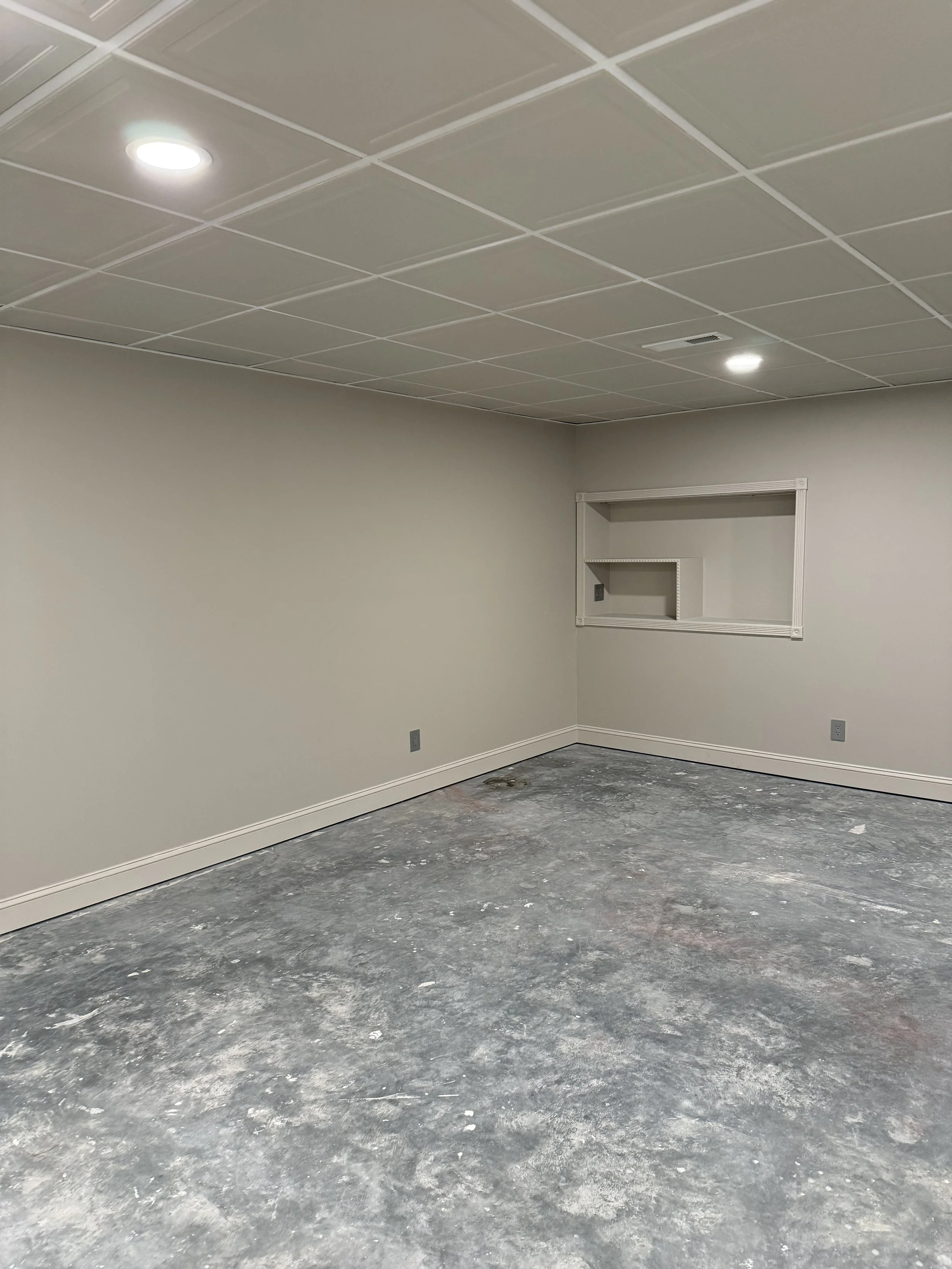 An empty room with beige walls, a white ceiling with square tiles, and a concrete floor. There is a small rectangular opening on the wall and two ceiling lights.