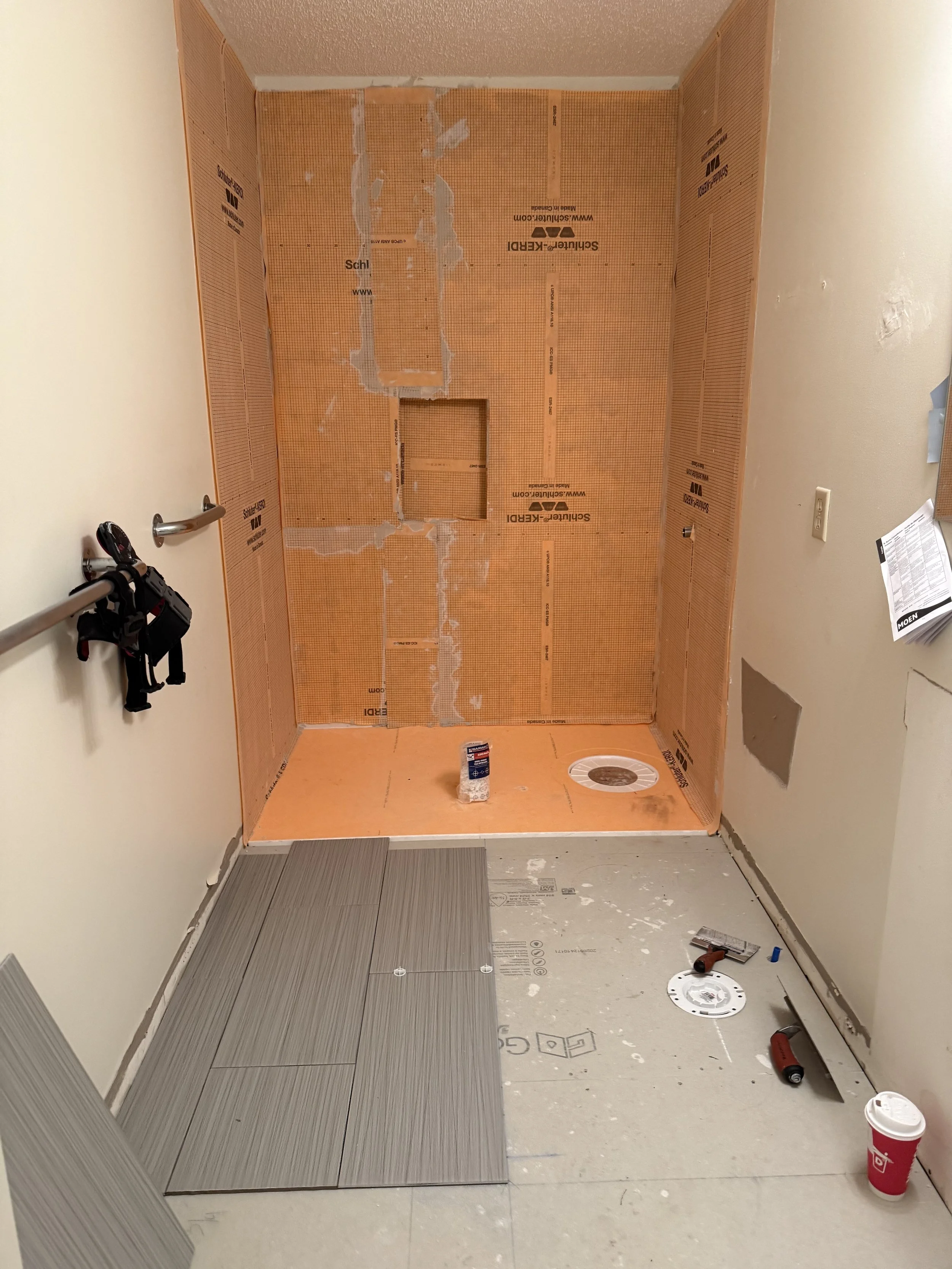 Bathroom under renovation with partially installed gray tiles on the floor, construction tools, and orange waterproofing membrane on the walls.