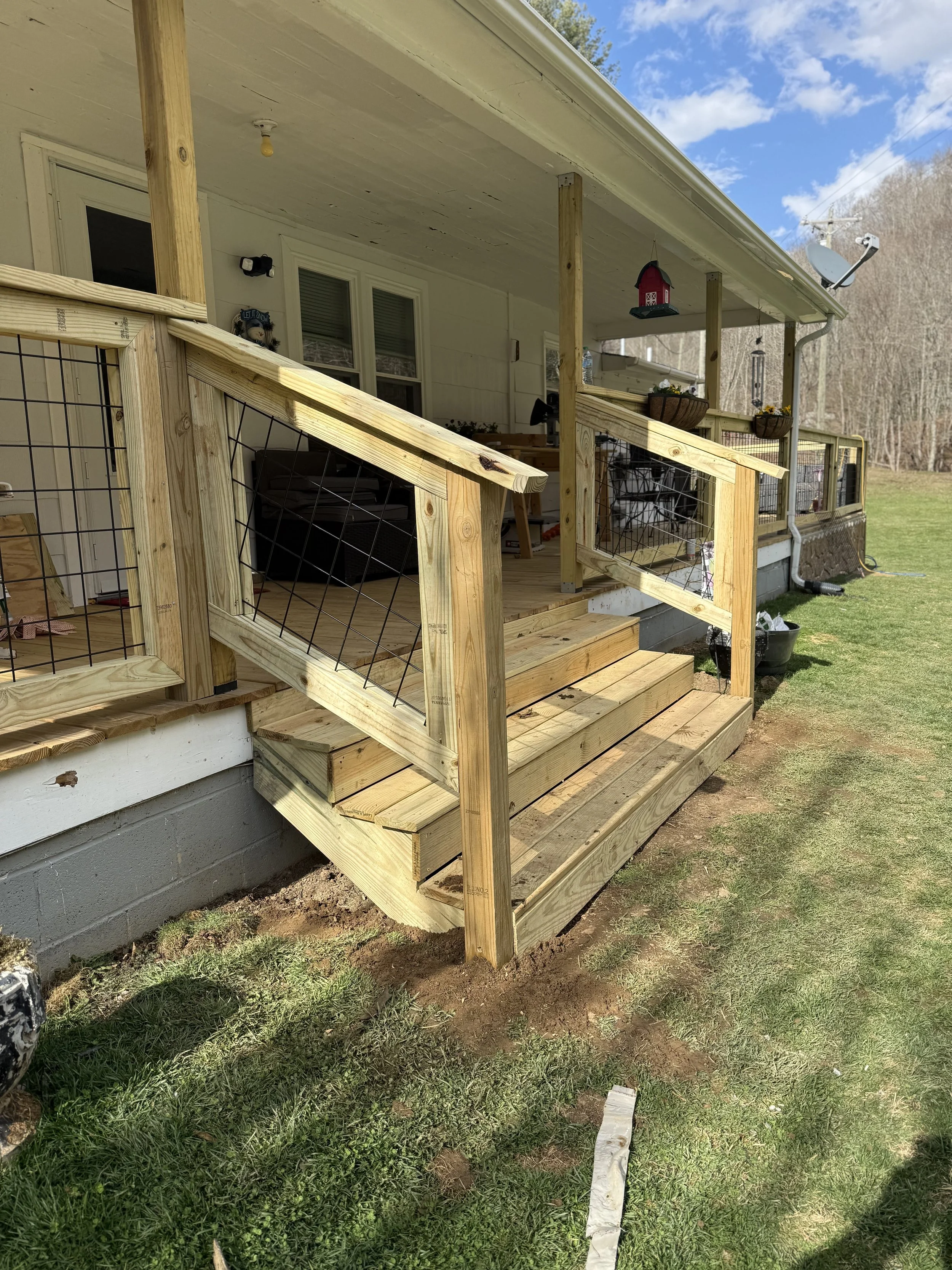 A house with a front porch under construction with new wooden stairs, railing, and support posts. The porch overlooks a grassy yard, and a satellite dish is mounted on the roof.