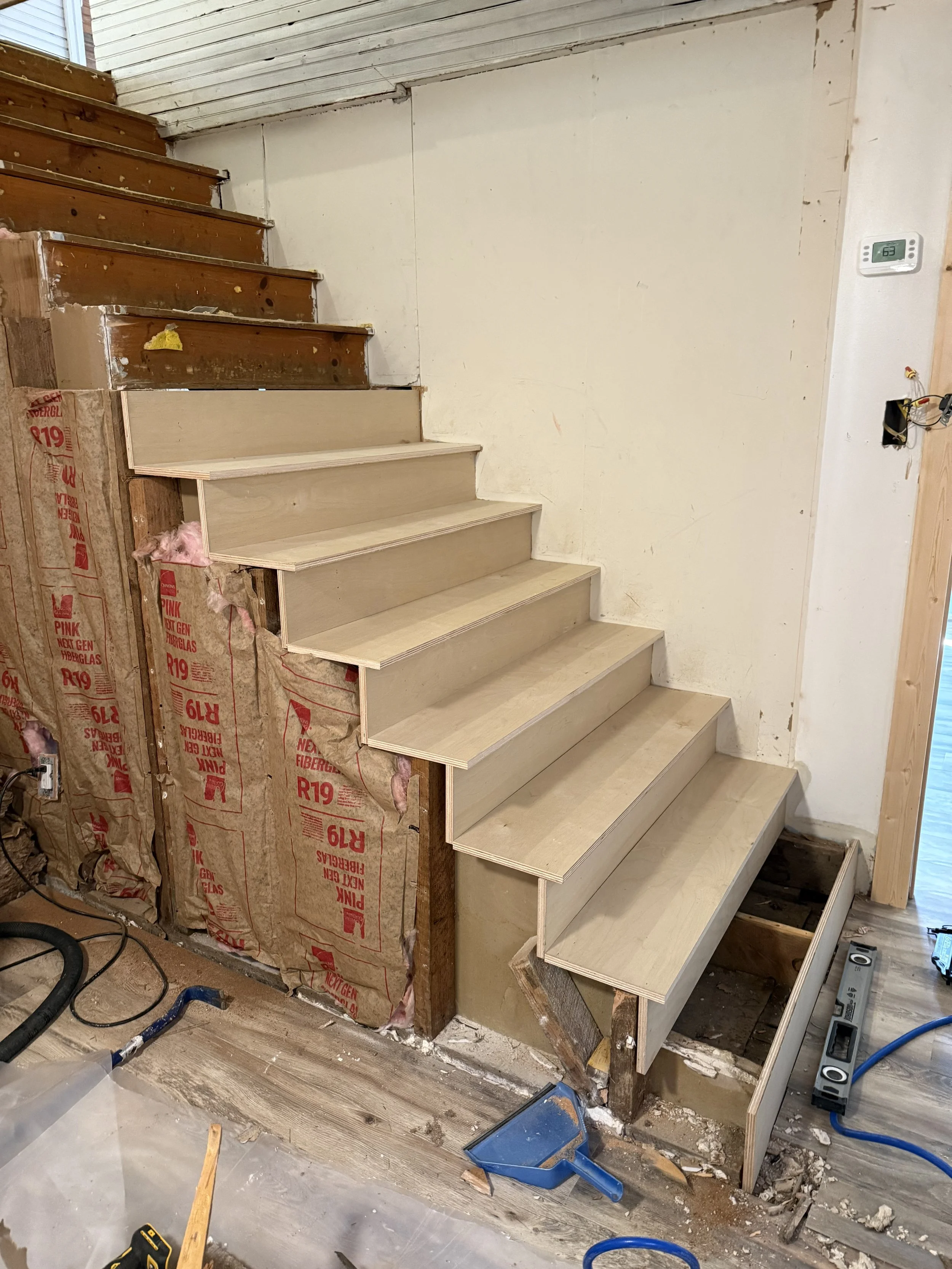 Inside a house renovation showing stairs under construction, with drywall partially installed, insulation exposed, and construction tools on the floor.