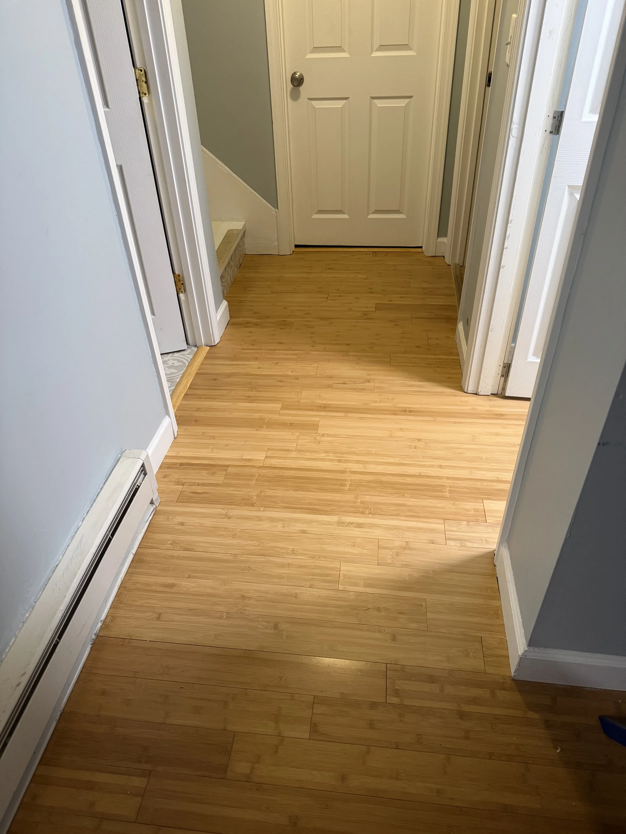 Wooden floor in a hallway with a closed white door at the end, a partial staircase to the left, and doorways on the right. Recently professionally installed by Appalachian Upgrades LLC