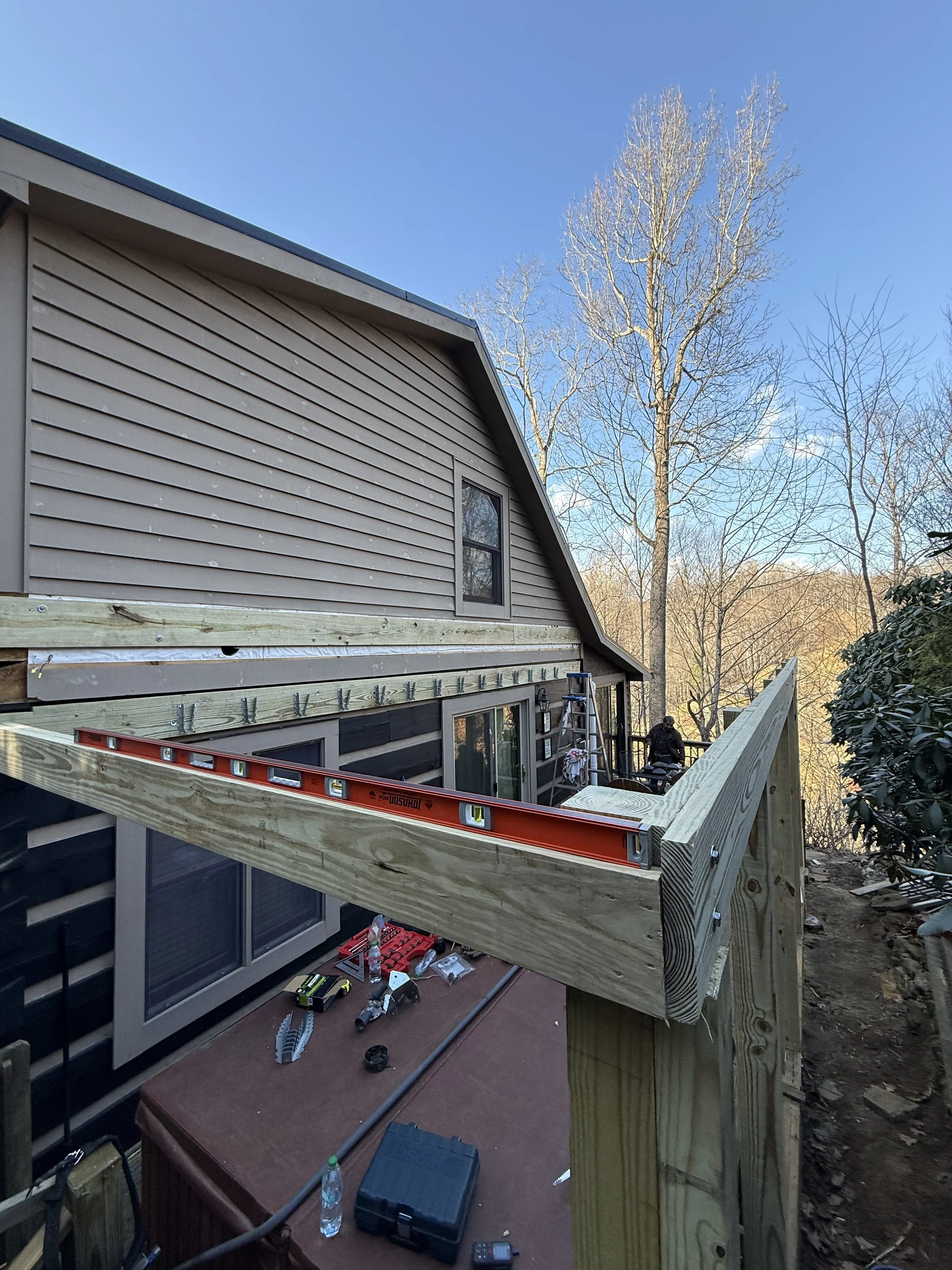 Construction of a wooden deck attached to a house with tools and equipment scattered on the deck, and a ladder leaning against the house.