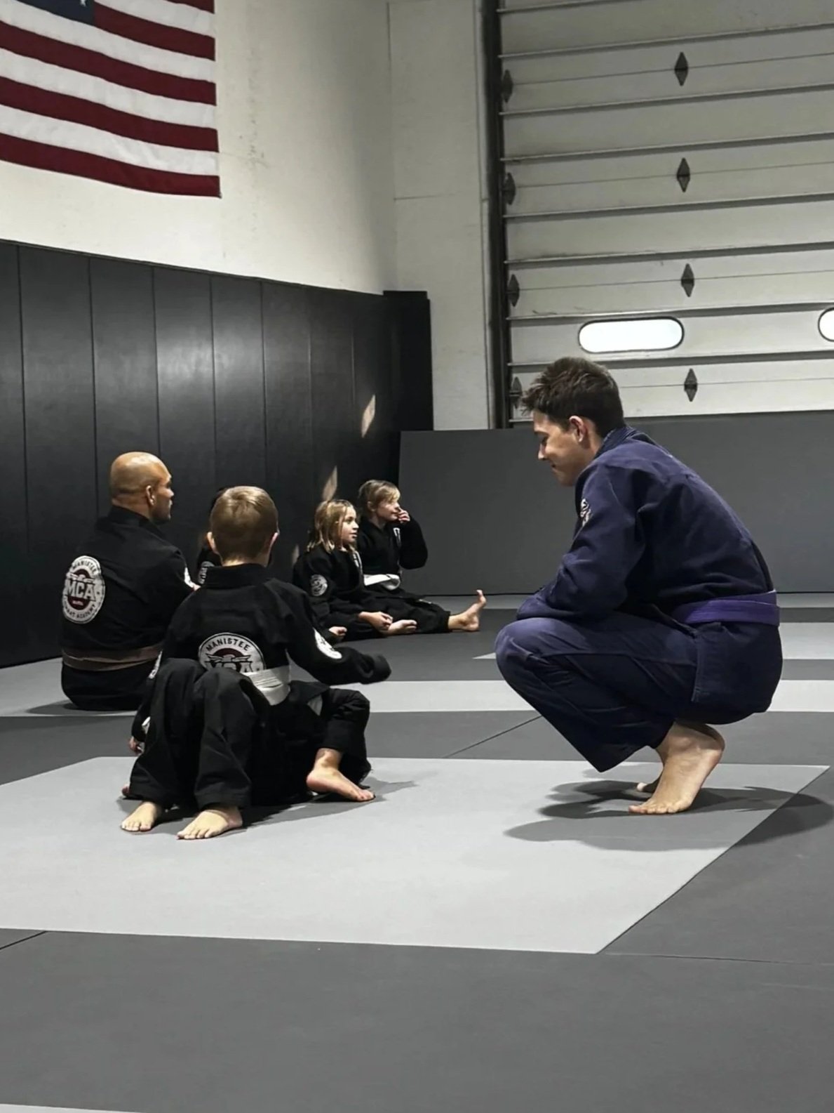 A martial arts instructor in a blue gi crouching in front of a group of children in black gis sitting cross-legged on martial arts mats inside a training gym.