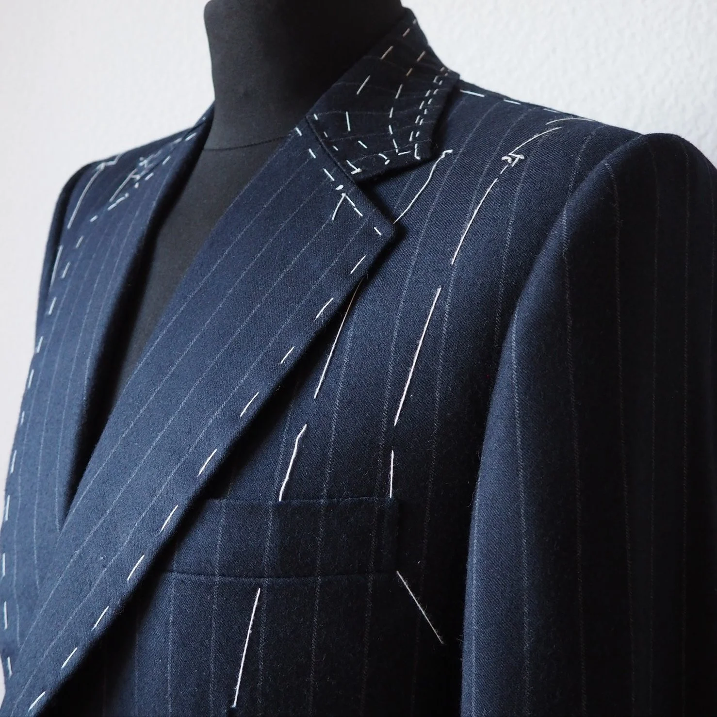 Close-up of a dark pinstripe suit jacket on a mannequin, with basting stitches visible, indicating it's an unfinished bespoke garment.
