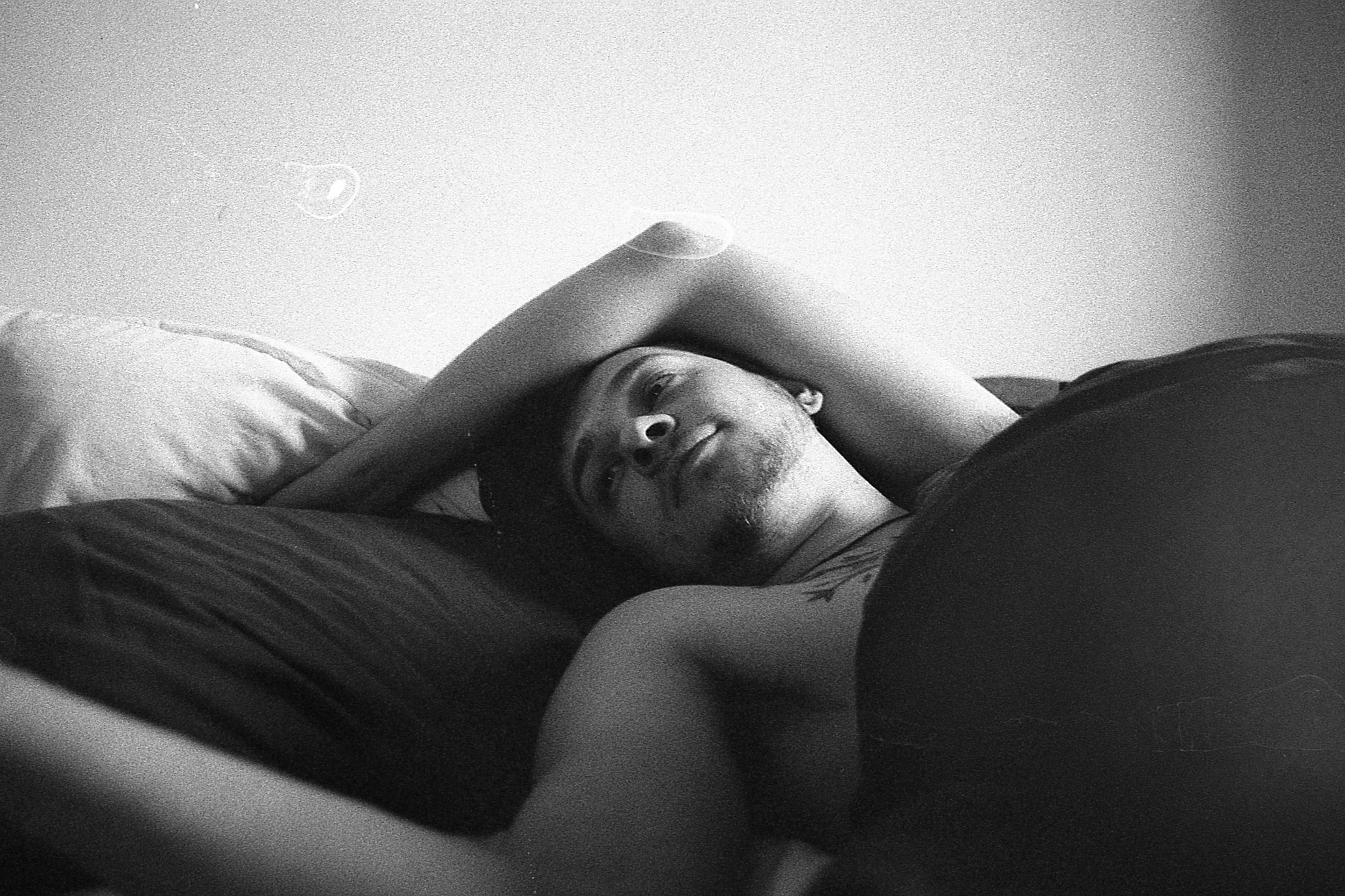 A man lying in bed, smiling, with one arm resting on his forehead, captured in black and white.