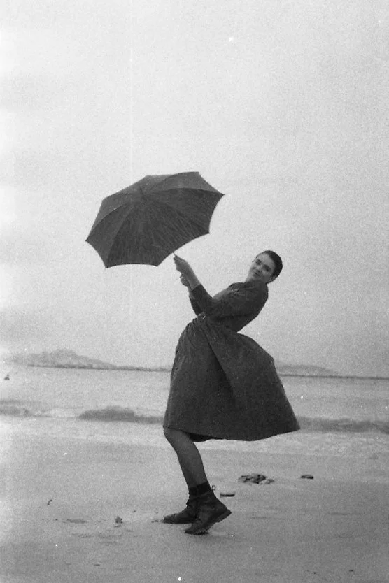 Woman standing on beach holding an umbrella, wearing a long coat and boots, with wind blowing her skirt
