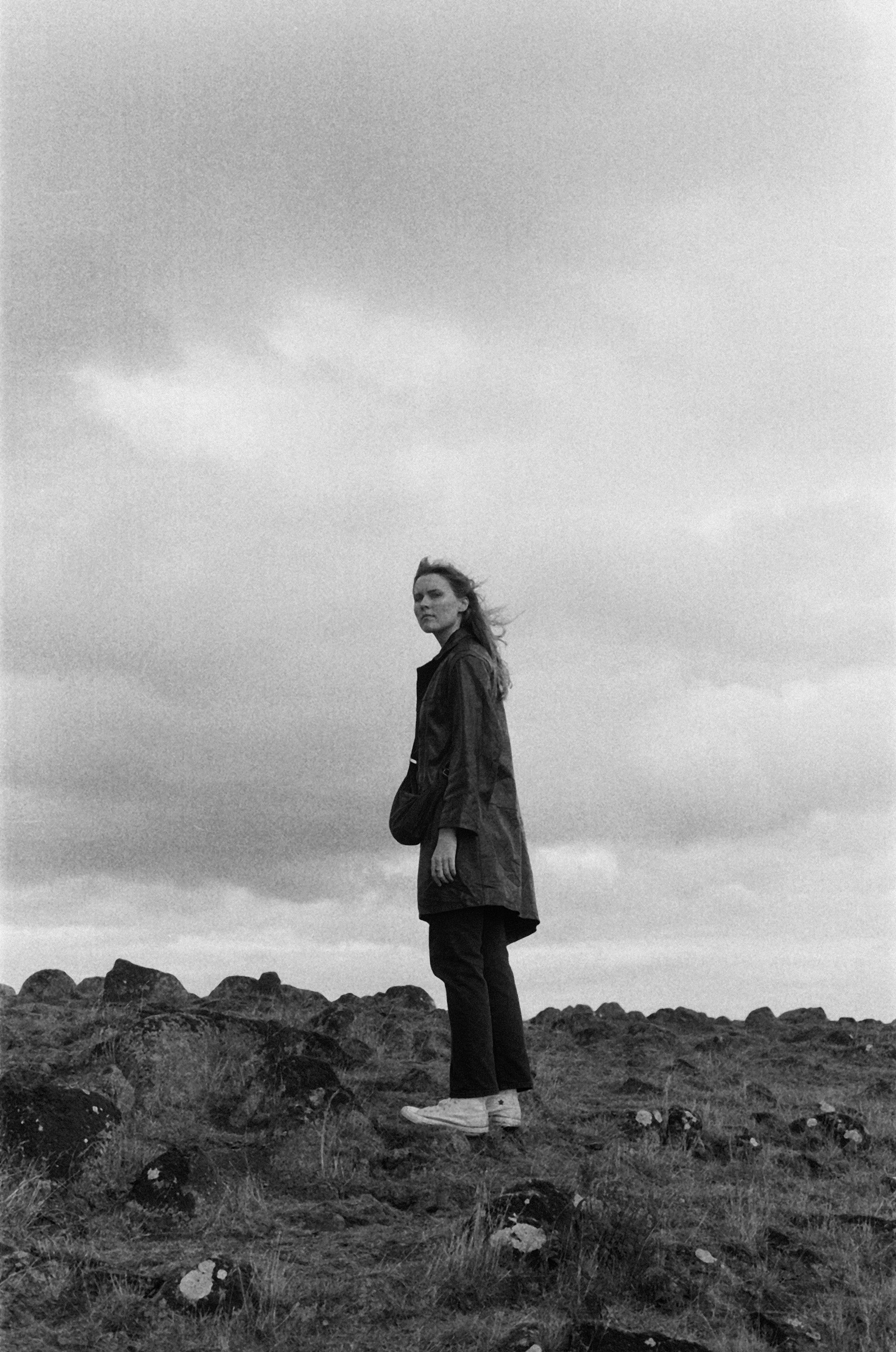 A woman standing on rocks in an open field with a cloudy sky in the background, dressed in a jacket, pants, and sneakers, captured in black and white.