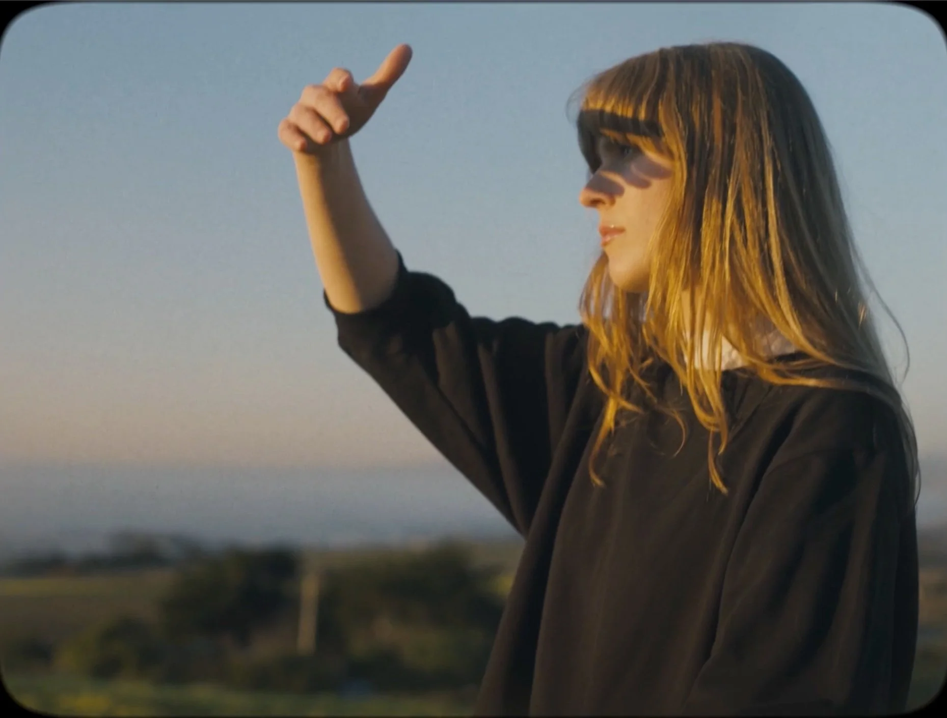 Lana Graves standing outdoors during sunset, with her arm raised and hand pointing upward, looking into the distance.