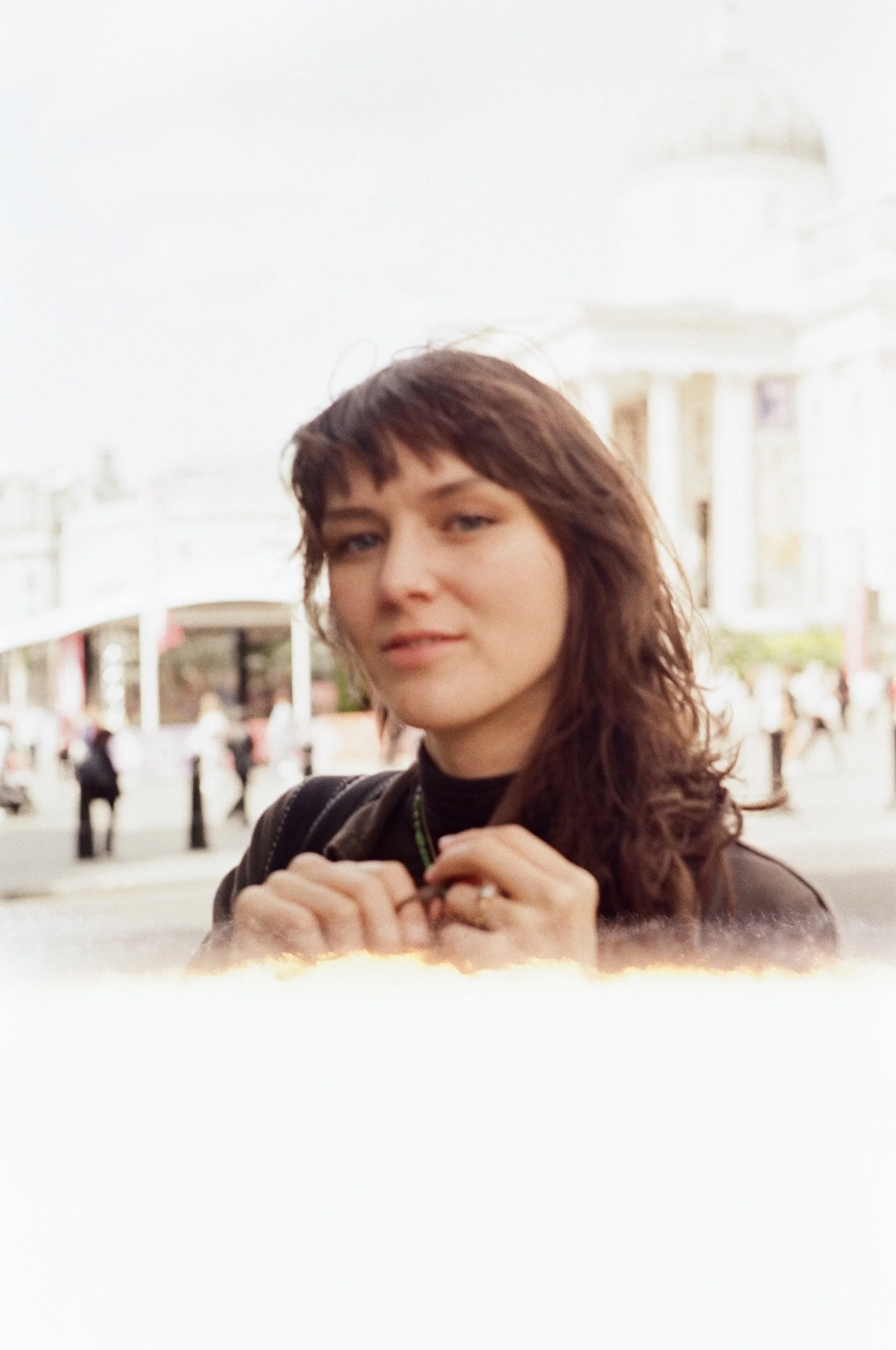 Bella Graves with brown hair and a black top standing outdoors in front of a white building with columns, holding her hands near her chest.