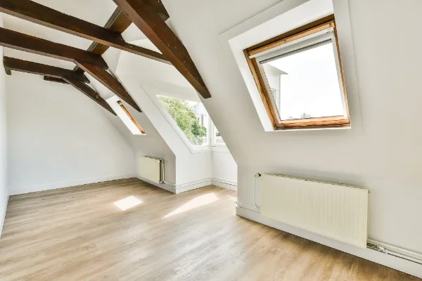 Empty attic room with white walls, wooden beams, and three skylight windows, sunlight on light wood floor.