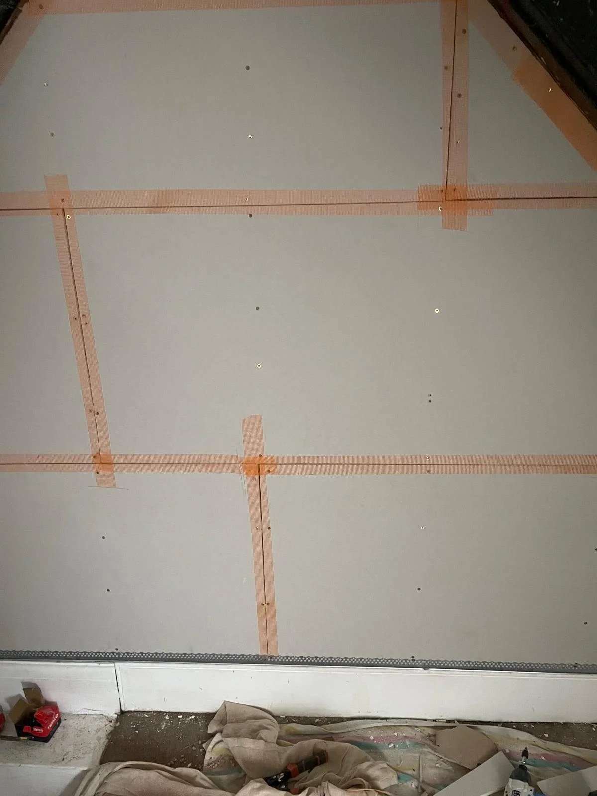 Ceiling drywall installation with exposed screws and orange tape, construction tools and materials on the floor.