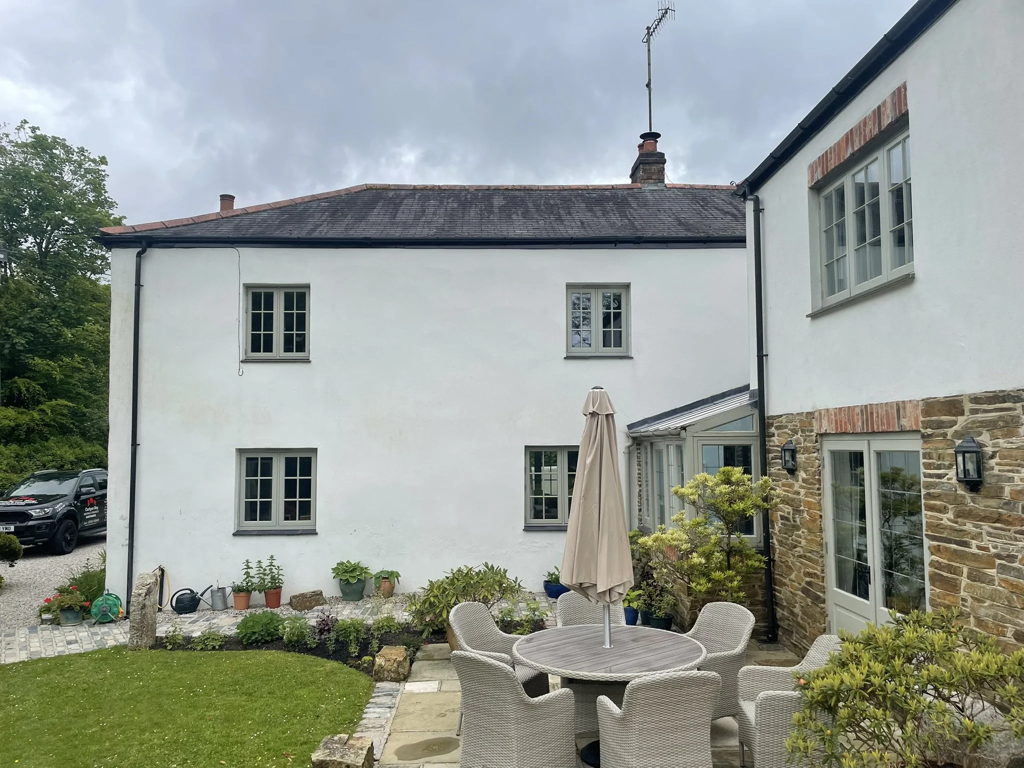 Backyard patio area with outdoor furniture, including a round table with an umbrella and several chairs, lush green lawn, various potted plants, and a white house with multiple windows and a brick section. Overcast sky.