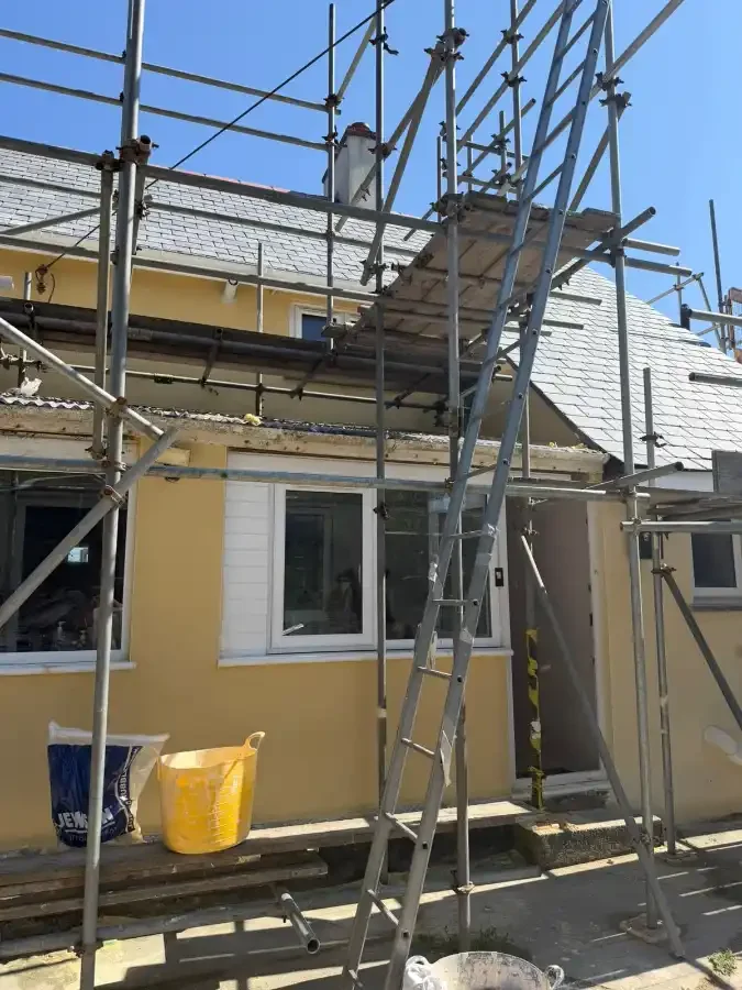 Construction scaffolding erected around a yellow house with a tiled roof, with a mountain visible in the background.