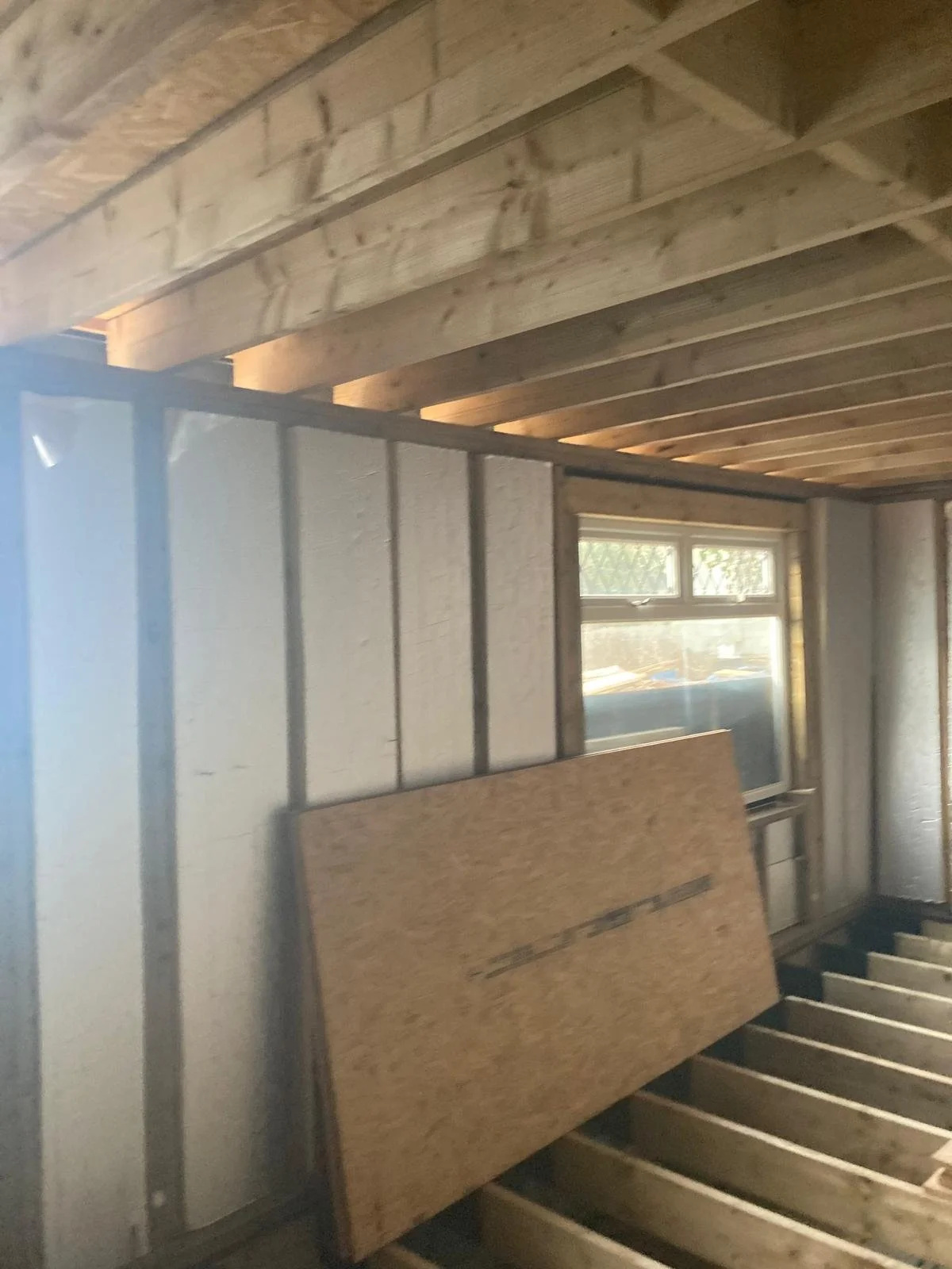 Interior of a room under construction with exposed wooden ceiling beams, a partially finished wall with metal framing, a window with a view of the outdoors, and a large piece of oriented strand board (OSB) leaning against the wall.