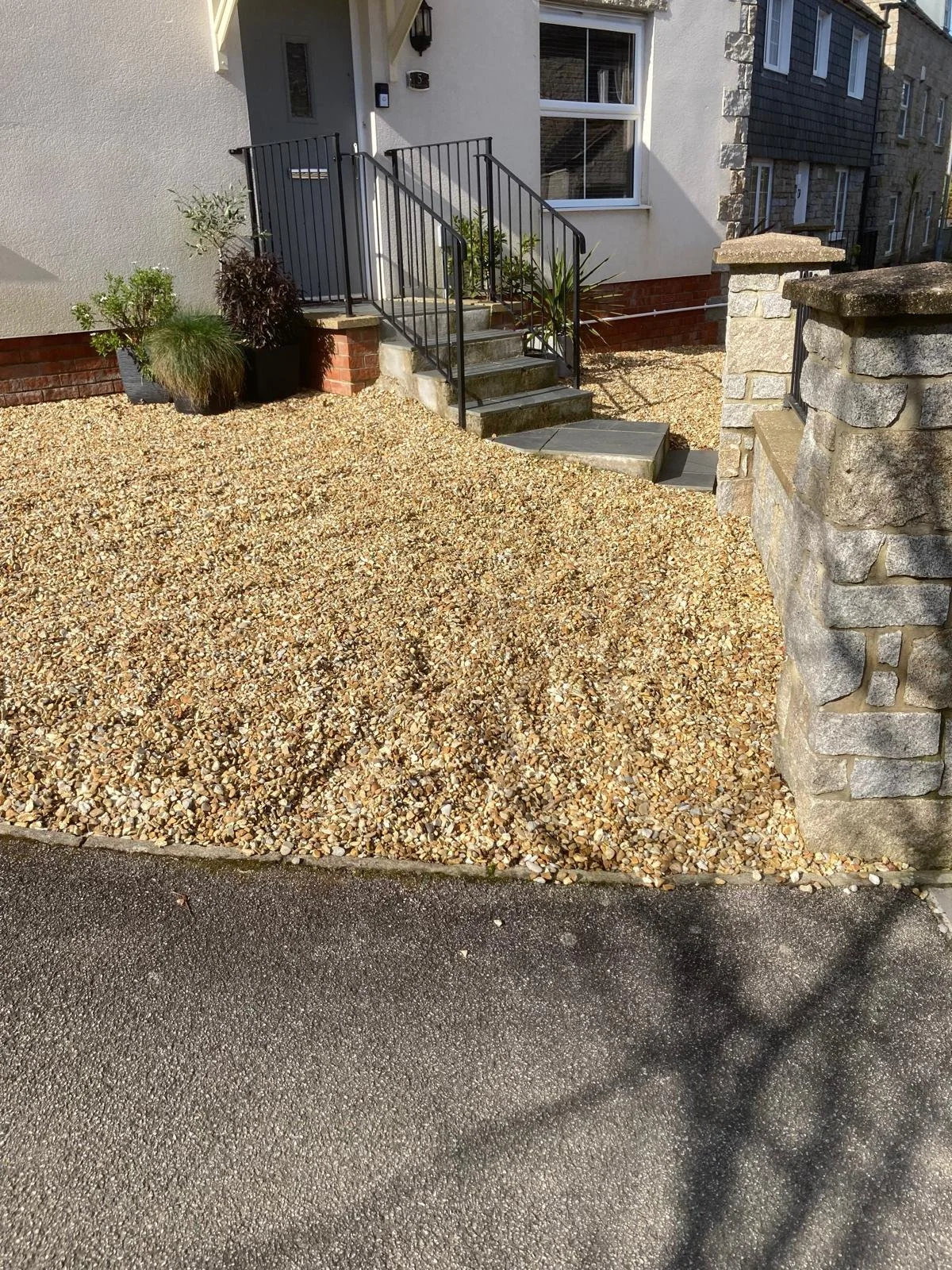 A gravel front yard with stairs leading to a house entrance, next to a stone pillar and a potted plant, with a paved driveway in the foreground.