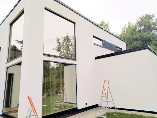 Modern white house with large windows, surrounded by greenery, and ladders standing in front of it.