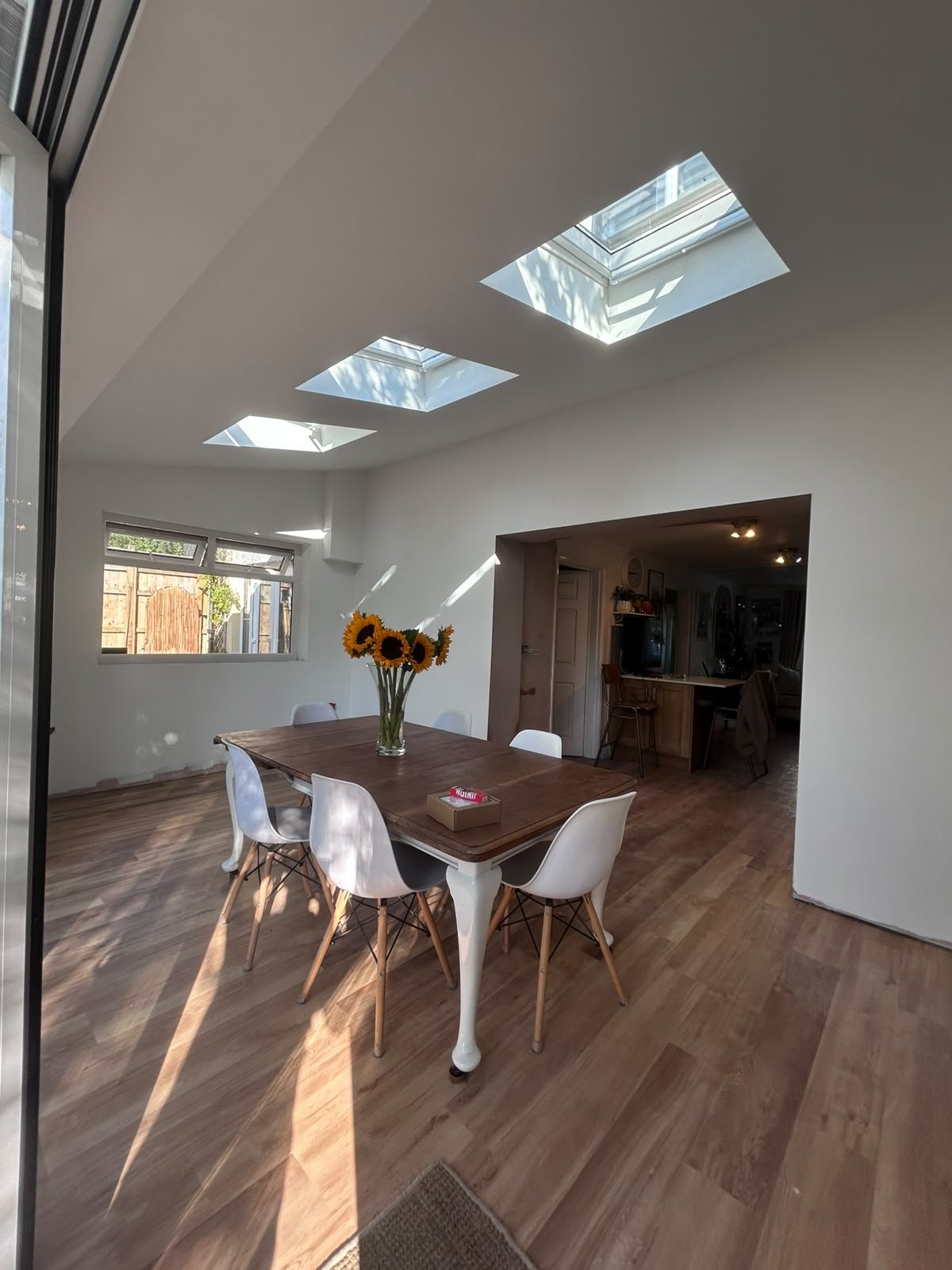 A dining room with a wooden table and six white chairs, a vase of sunflowers, and skylights letting in natural light.