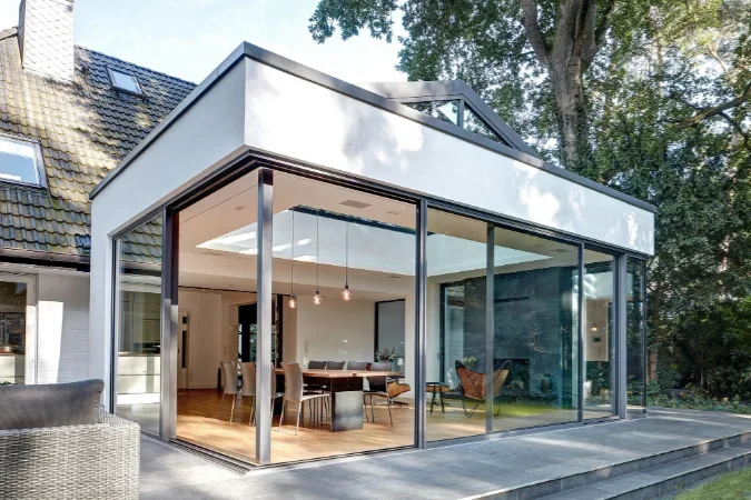 Modern glass house with a dining area and outdoor patio, surrounded by trees.