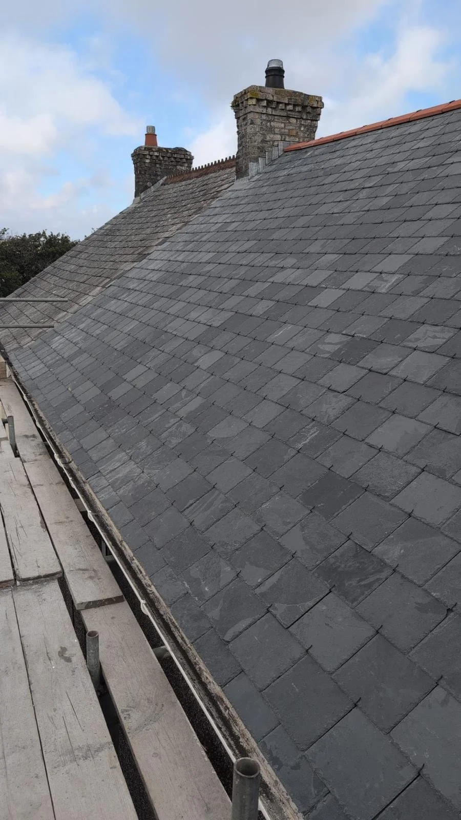 Rooftop covered with gray slate tiles, two brick chimneys, blue sky with clouds, part of a wooden scaffolding on the left side.