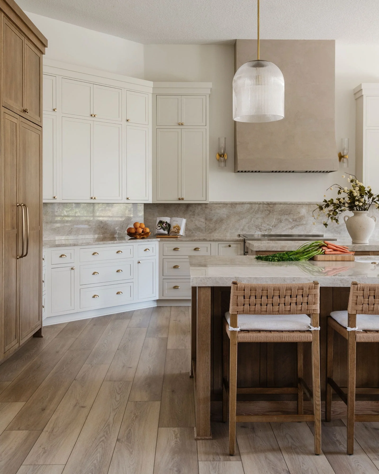 Warm neutral kitchen with white cabinetry, wood accents, stone countertops, a large island with woven barstools, and a plaster-style range hood.