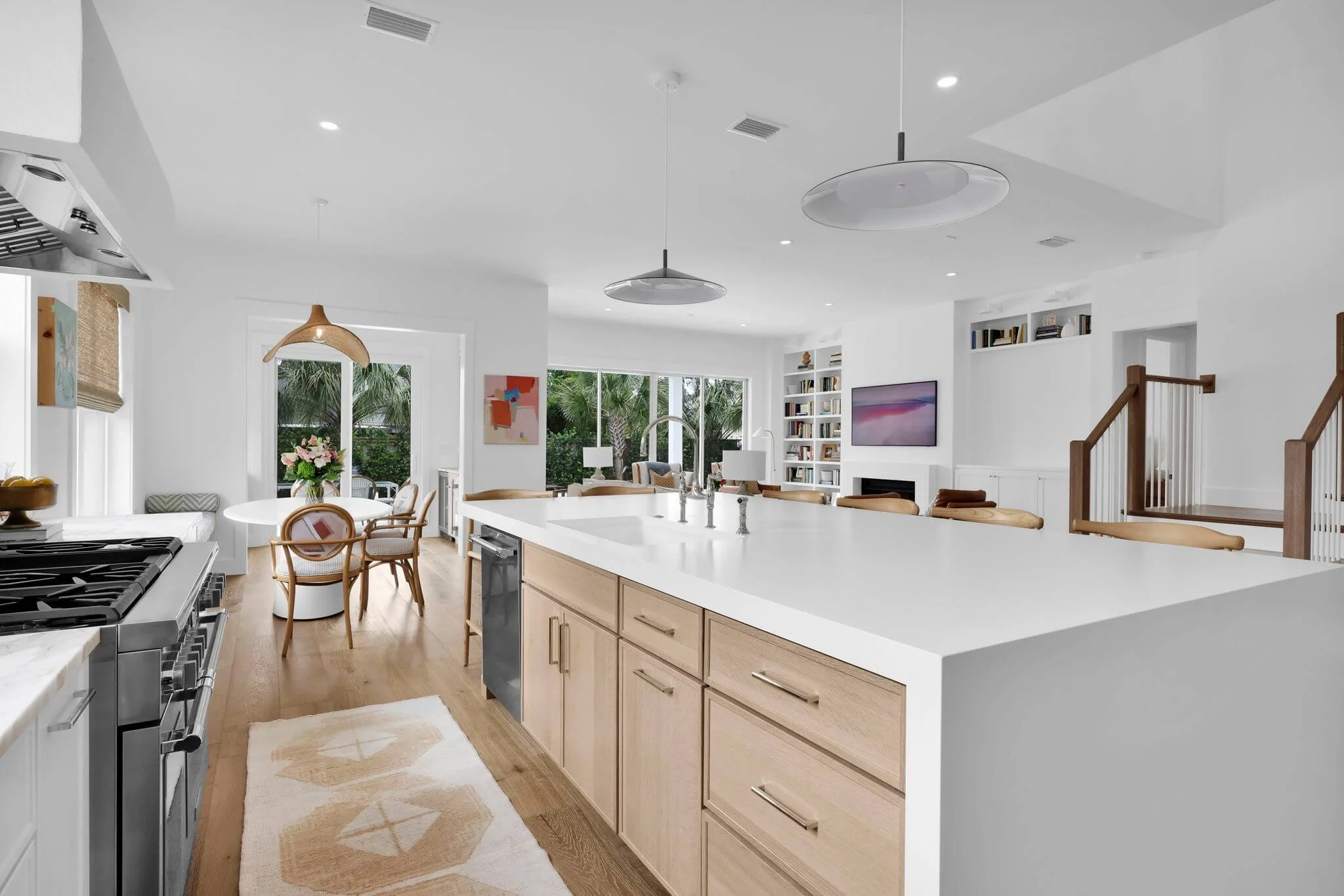 Bright open-concept kitchen and living space featuring a large white island with light wood cabinetry, modern pendant lights, a round dining table with floral centerpiece, and full-height windows overlooking palm trees.