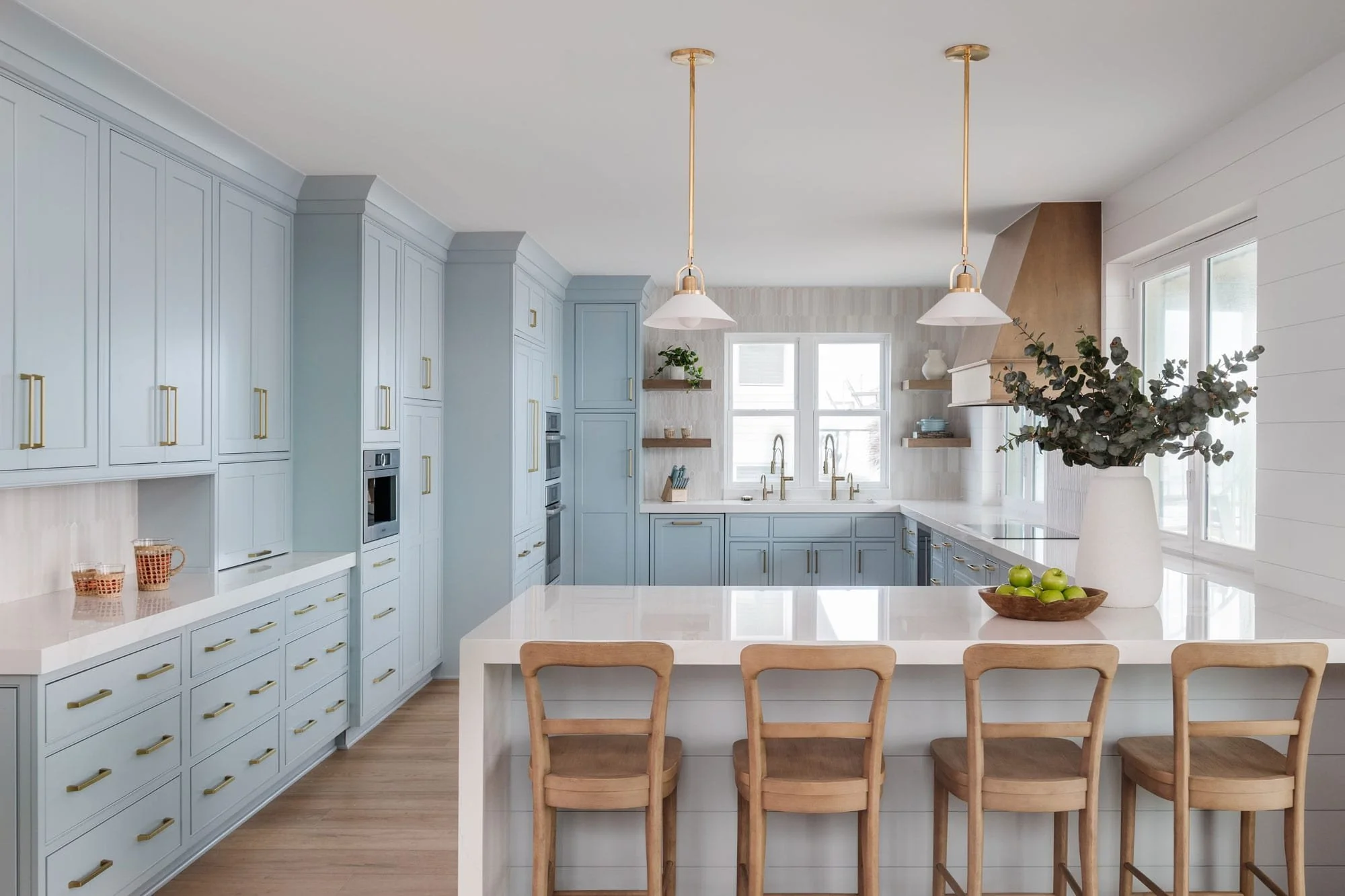 Light blue kitchen with shaker cabinets, white quartz island, brass hardware, pendant lights, and wood barstools.