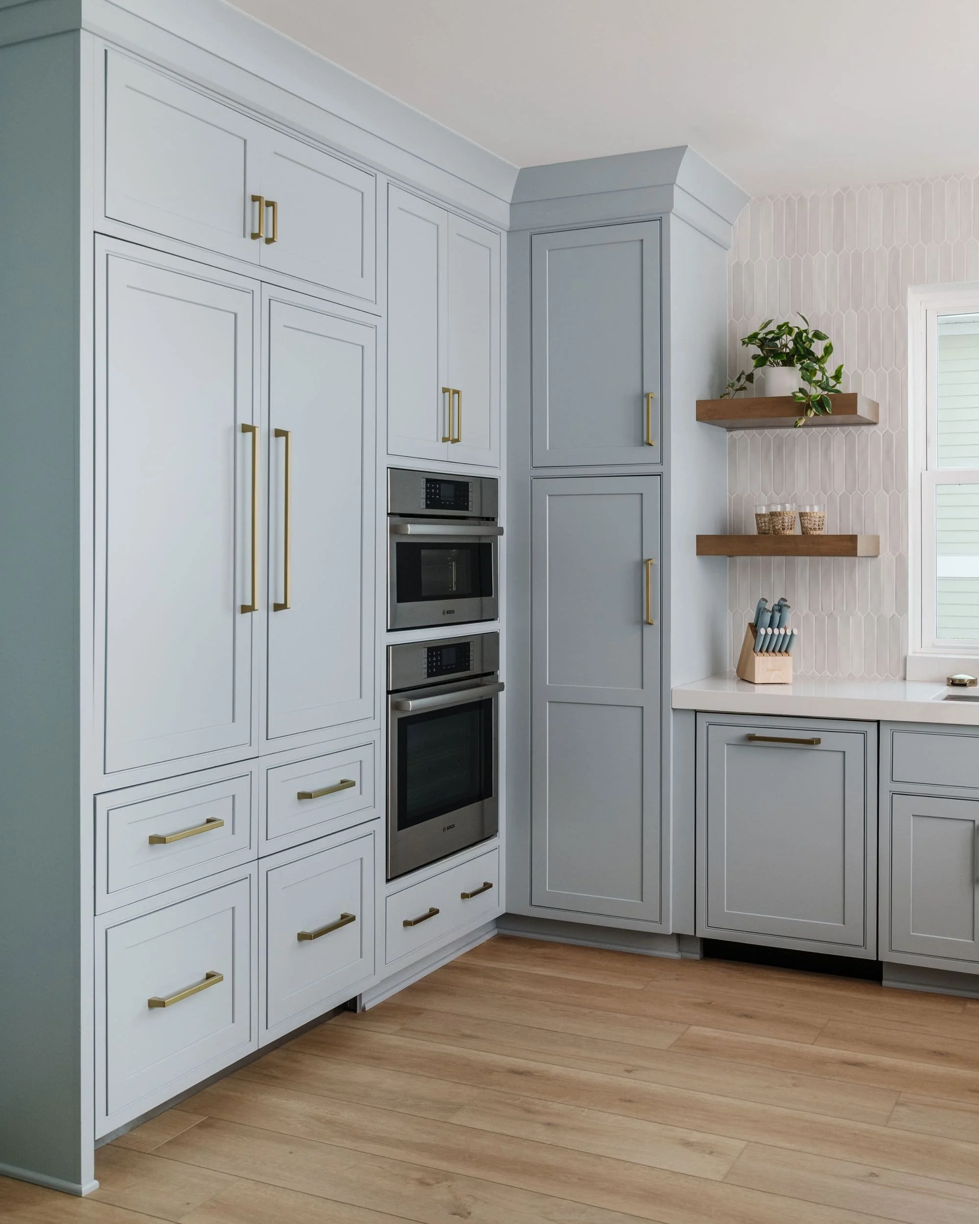 Corner view of a kitchen with built-in appliances and soft blue custom cabinetry featuring brass hardware, wood floating shelves, light oak floors, and a white textured backsplash.