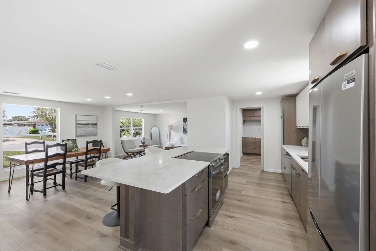 Open-concept kitchen with large island cooktop, light countertops, wood cabinetry, stainless steel appliances, and adjoining dining and living areas.