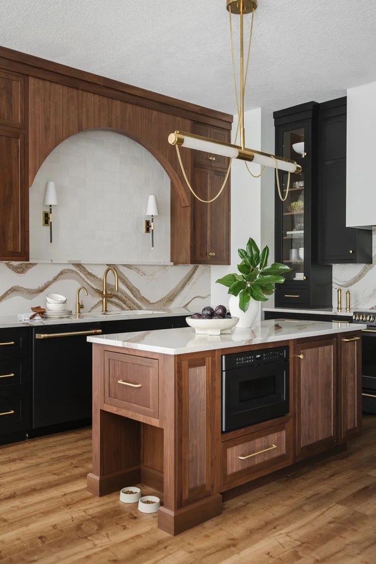 Kitchen with rich wood cabinetry, black accents, stone countertops, arched hood detail, and brass lighting over an island.