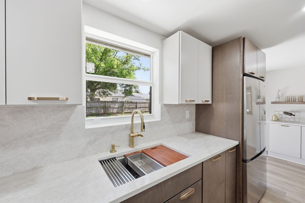 Modern kitchen sink area with brass faucet, integrated accessories, light countertops, and wood cabinetry.