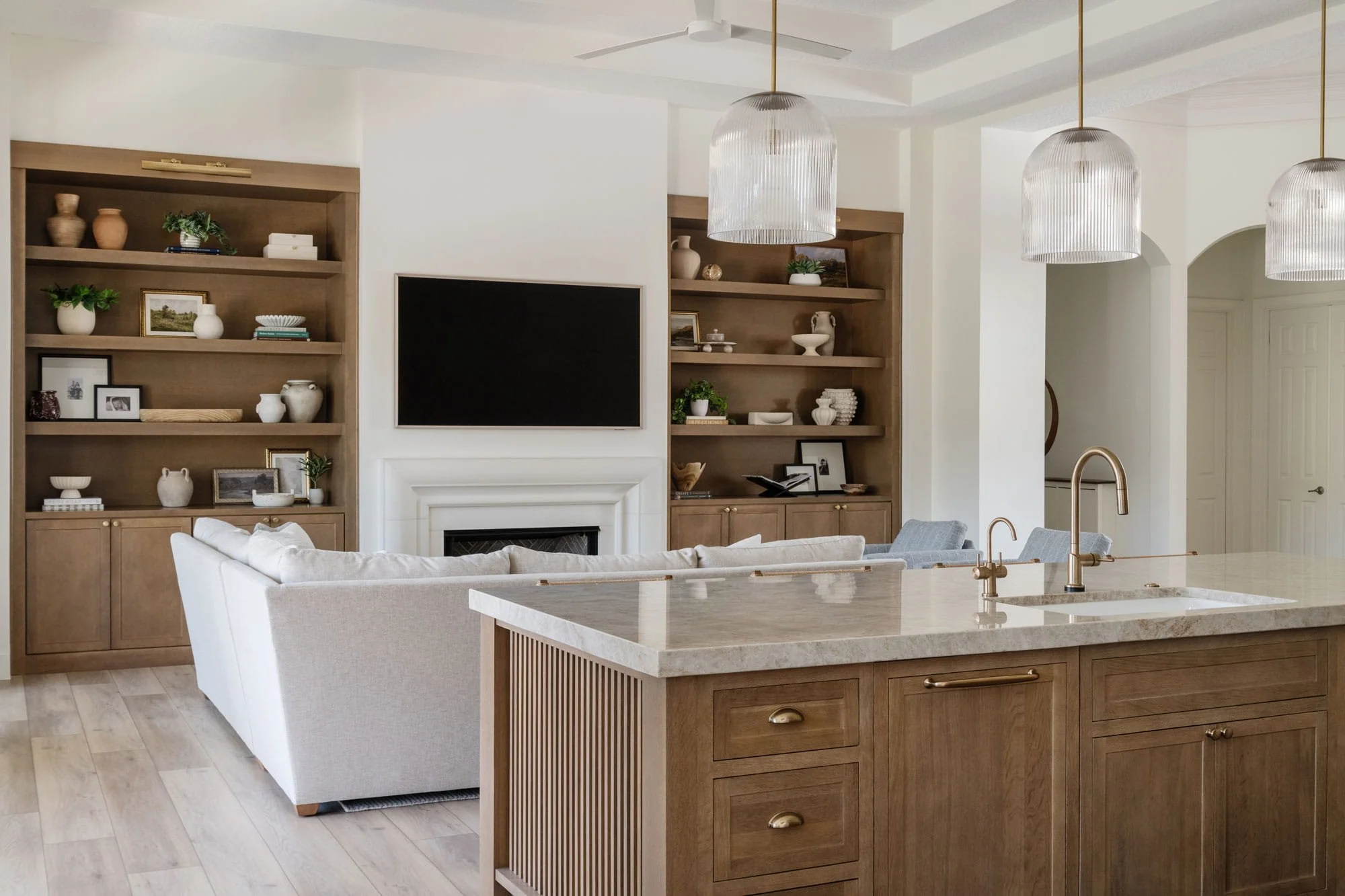 Open-concept kitchen and living room with wood cabinetry, stone island, brass fixtures, and built-in shelving around a fireplace and TV.