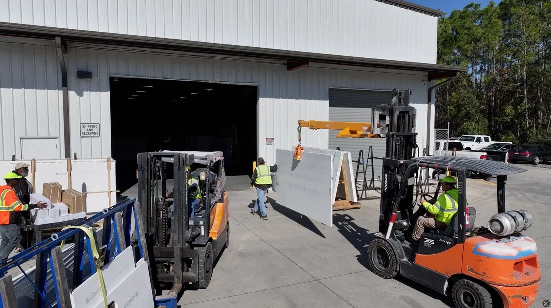 Workers using forklifts to move large stone slabs outside a warehouse during countertop fabrication and shipping operations.