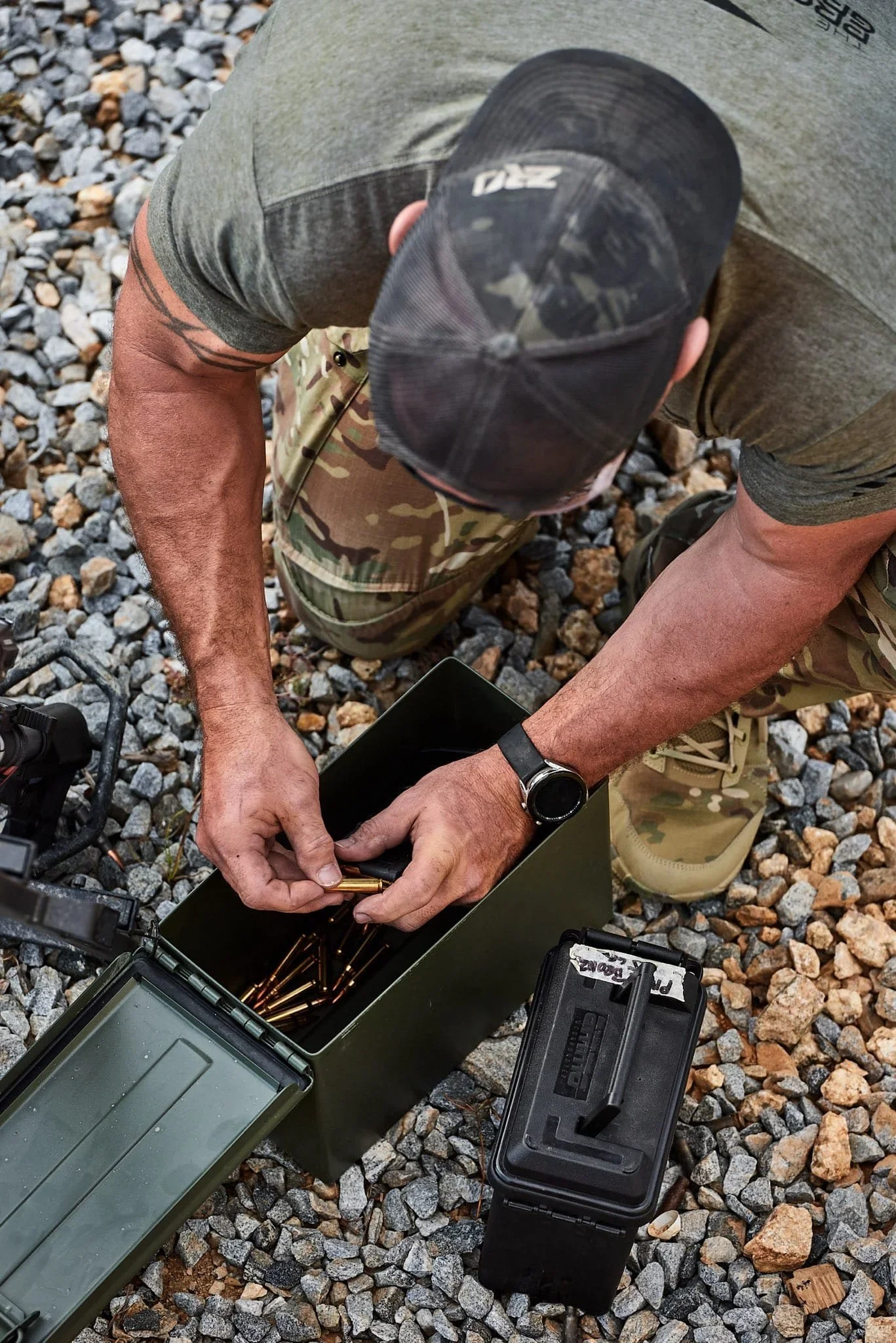 A man wearing camouflage pants, a gray T-shirt, and a baseball cap is kneeling on a gravel surface, loading brass ammunition cartridges into a green metal box. There is a black ammunition case nearby, and a camera is partially visible in the lower left corner.