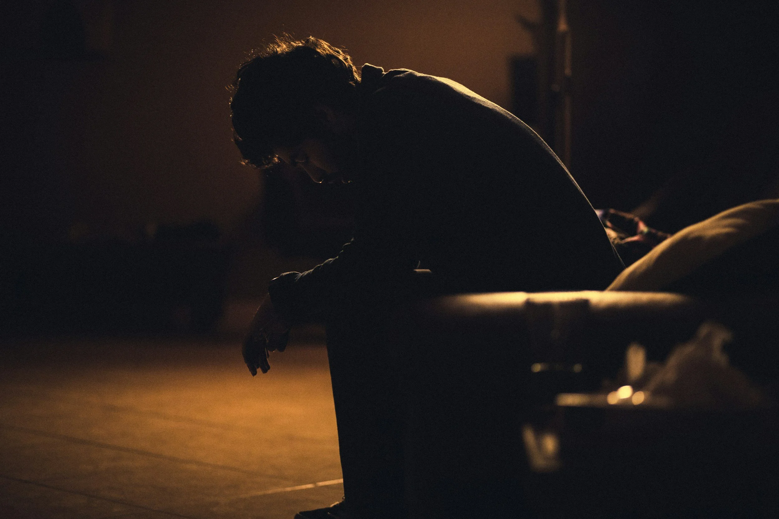 Silhouette of a person sitting on a bench in a dimly lit room, with head bowed and arm resting on knees.