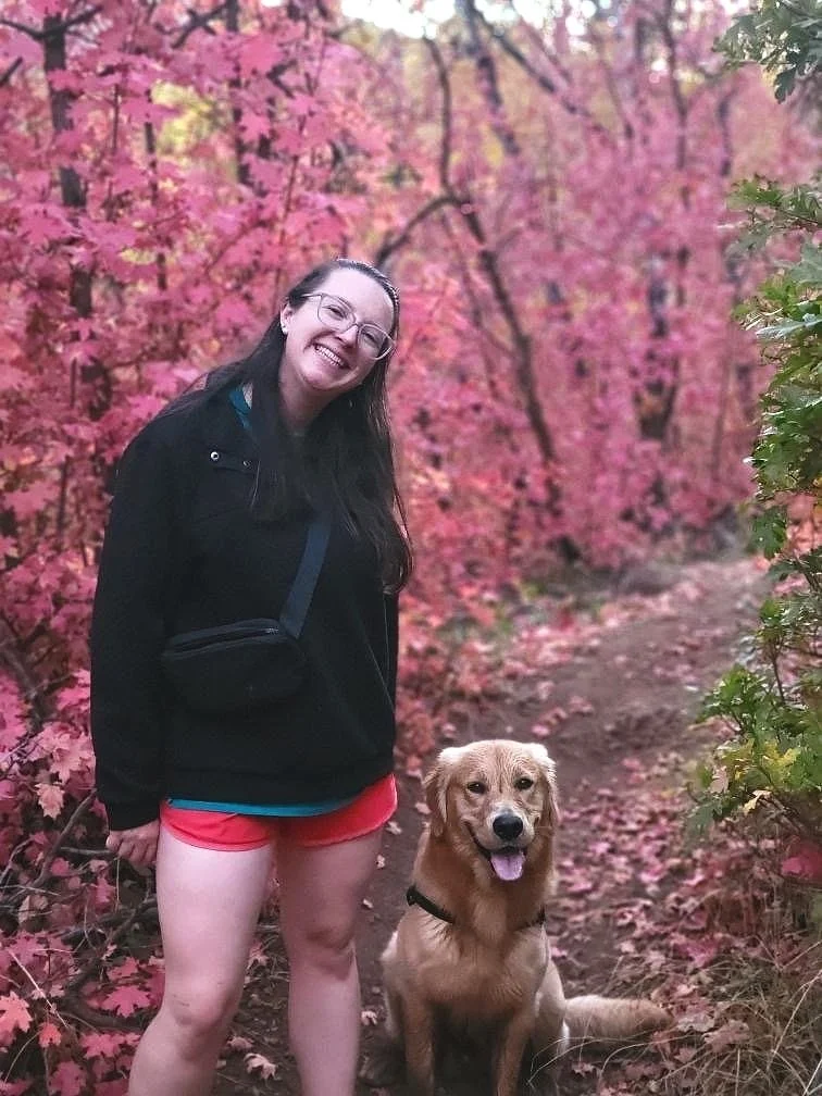 A woman smiling in a black jacket and pink shorts standing next to a golden retriever in a forest with pink autumn leaves on trees.