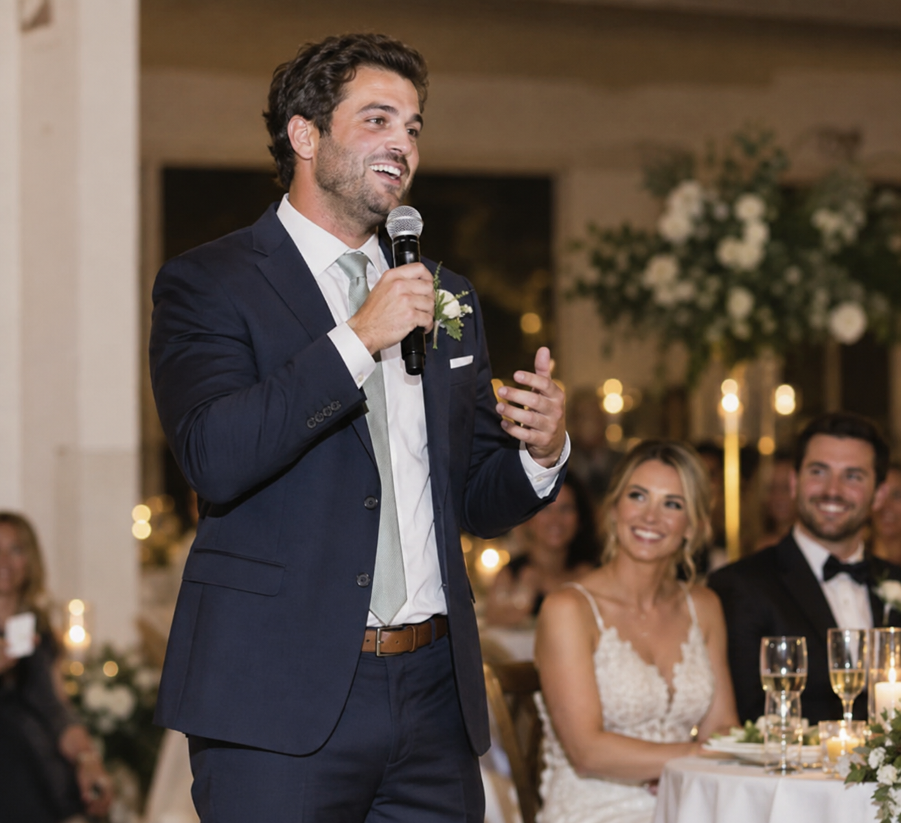 A man in a navy suit giving a speech at a wedding reception, holding a microphone, with a smiling woman in a white wedding dress and a man in a tuxedo sitting at the table.