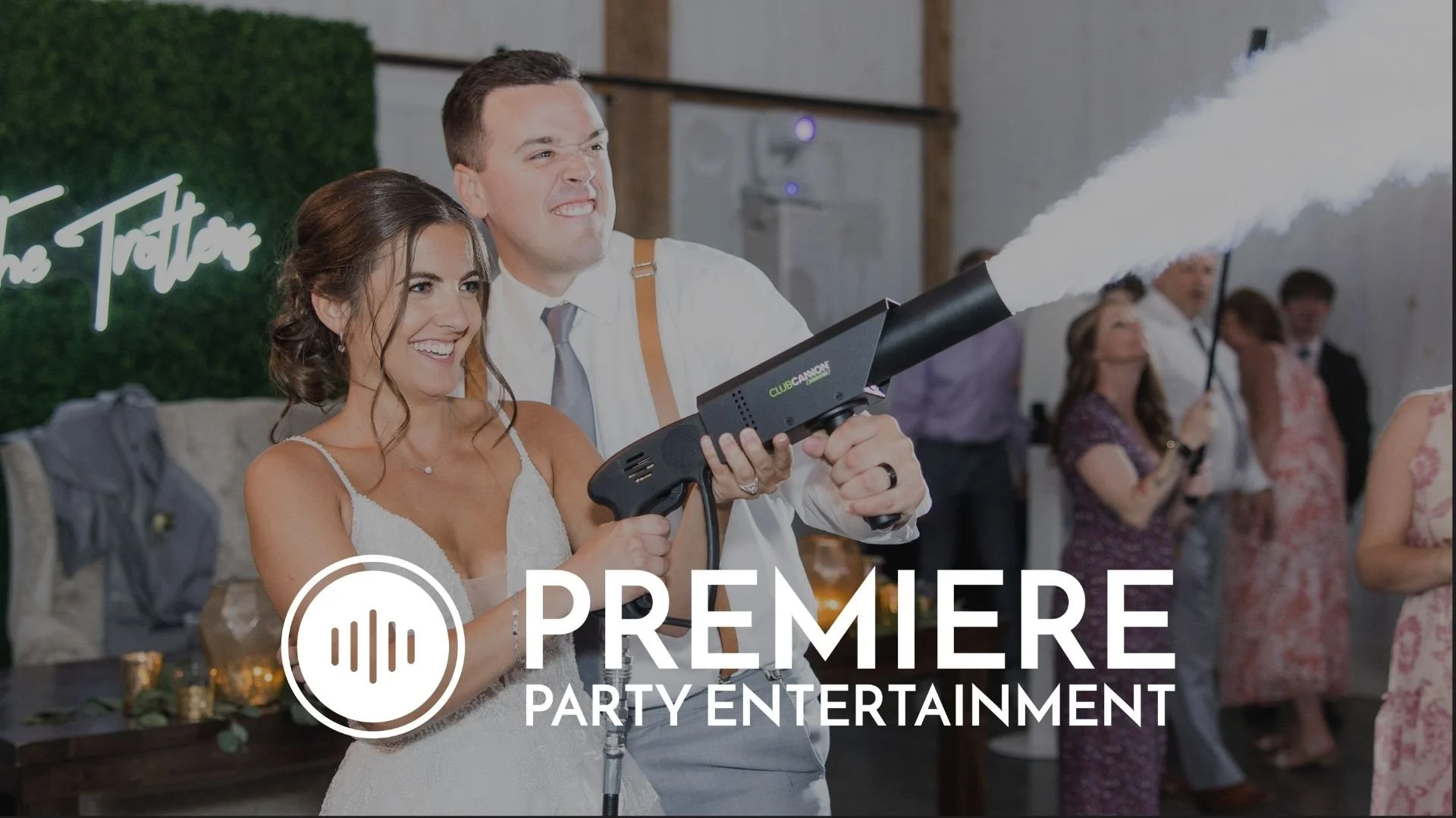 A man and woman at a wedding reception using a fog machine with a party crowd in the background.