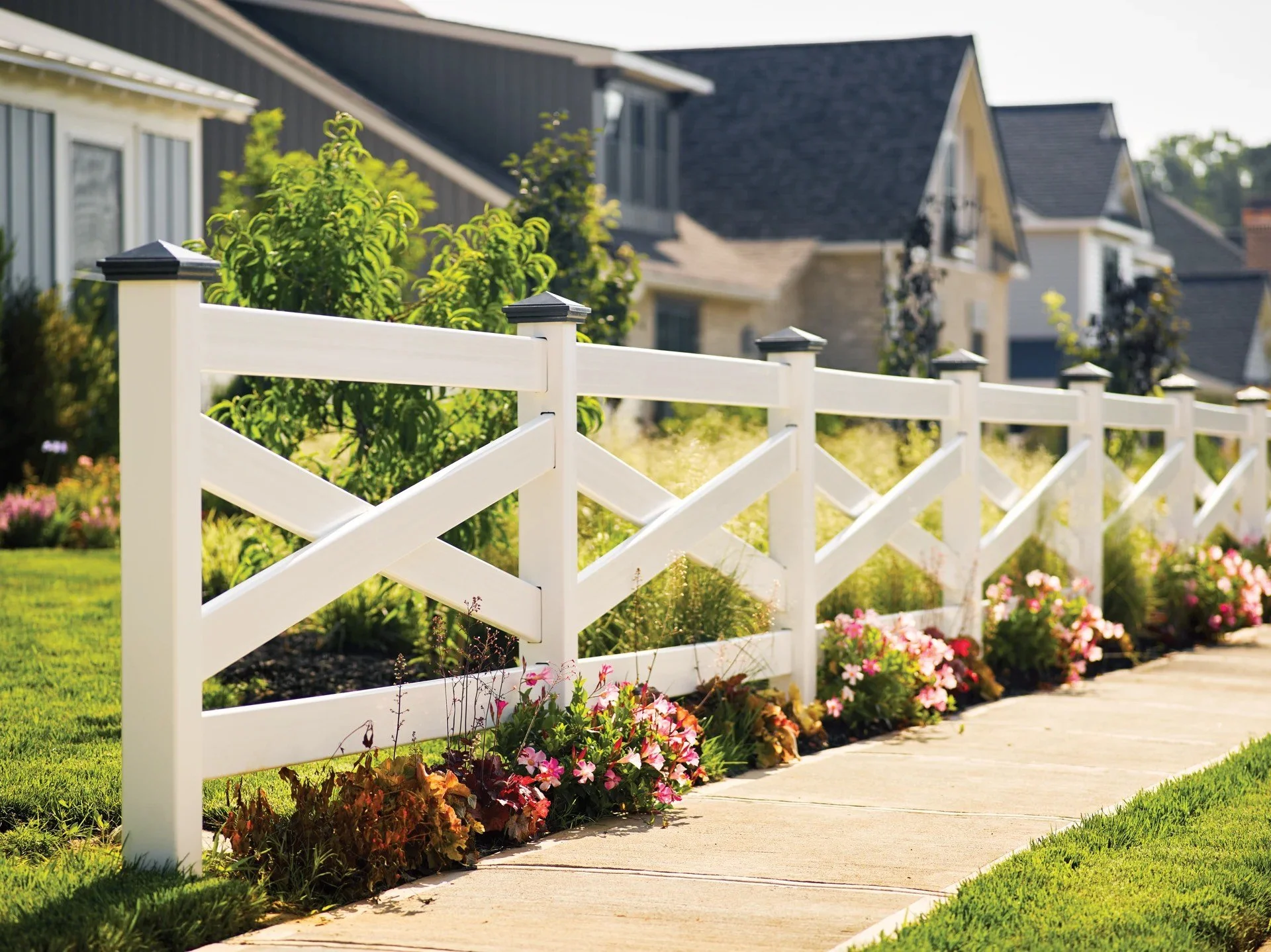 White picket fence with flowers and green lawn, houses in background, sunny day.