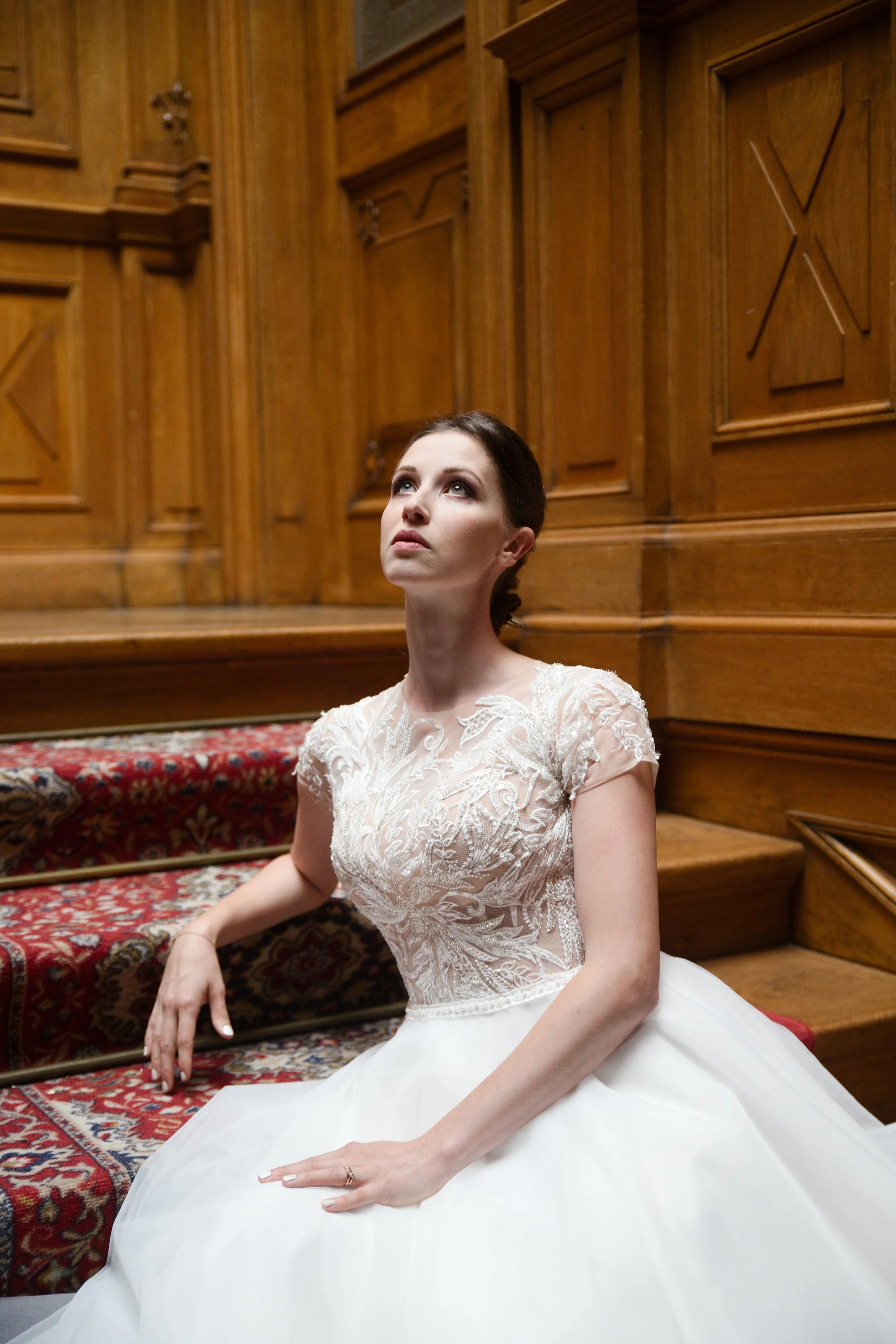 Bride in white lace gown seated on ornate red carpeted stairs with wood paneling background.