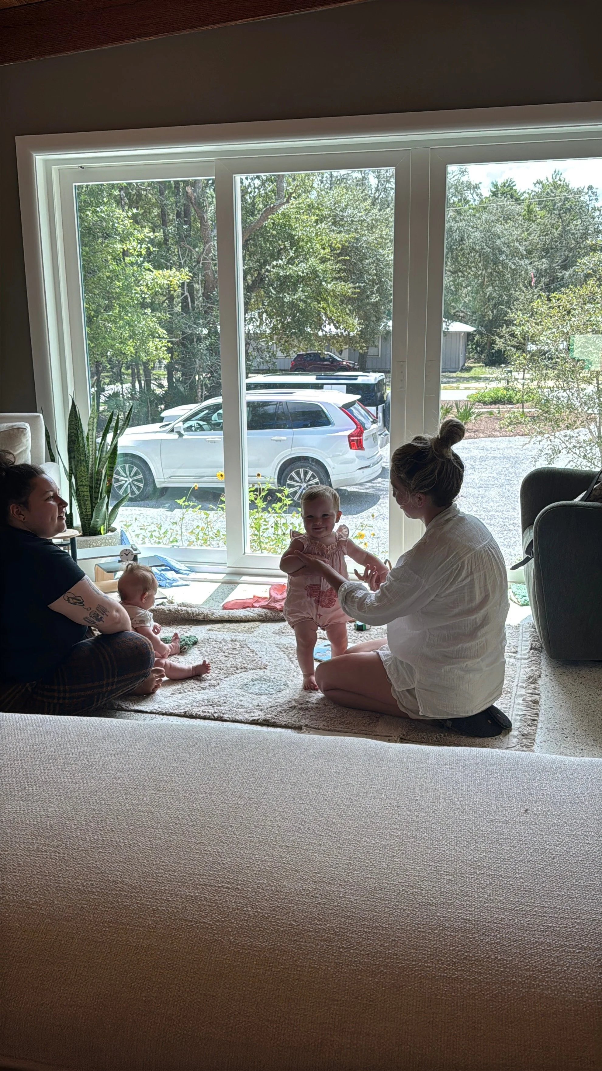 A woman and two children sitting on a carpet in front of a large window, with a view of cars outside. One child is standing while the other is sitting on the floor, and the woman appears to be helping the standing child.