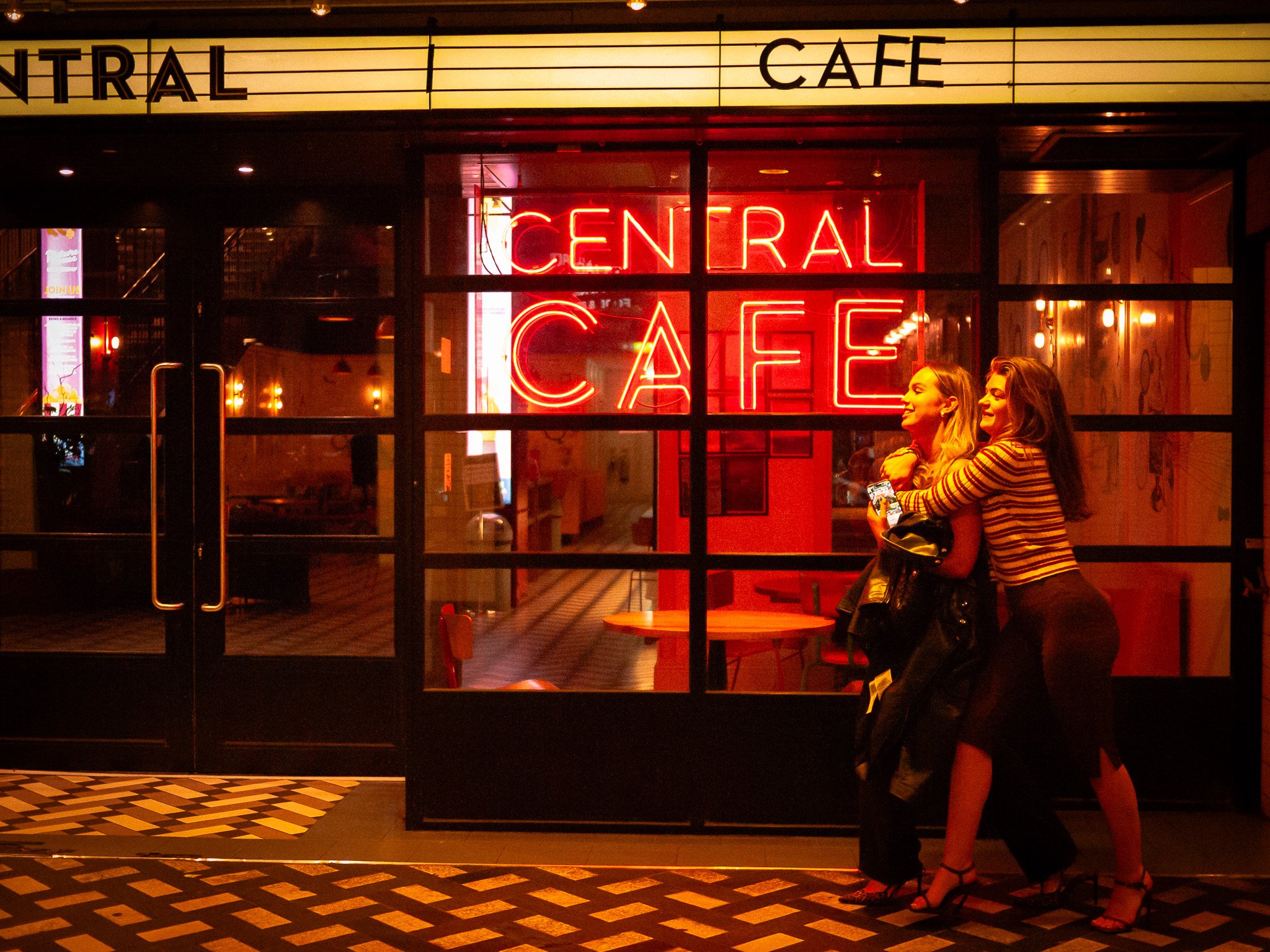 Two women hugging outside a cafe with a bright red neon sign that says 'Central Cafe' at night.
