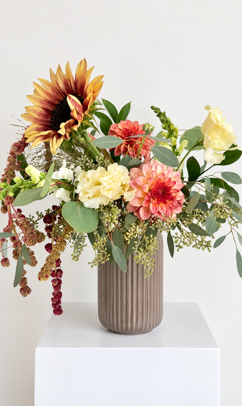 Elegant floral arrangement in a tall, ribbed ceramic vase with sunflowers, dahlias, carnations, and assorted greenery on a white pedestal against a neutral background.